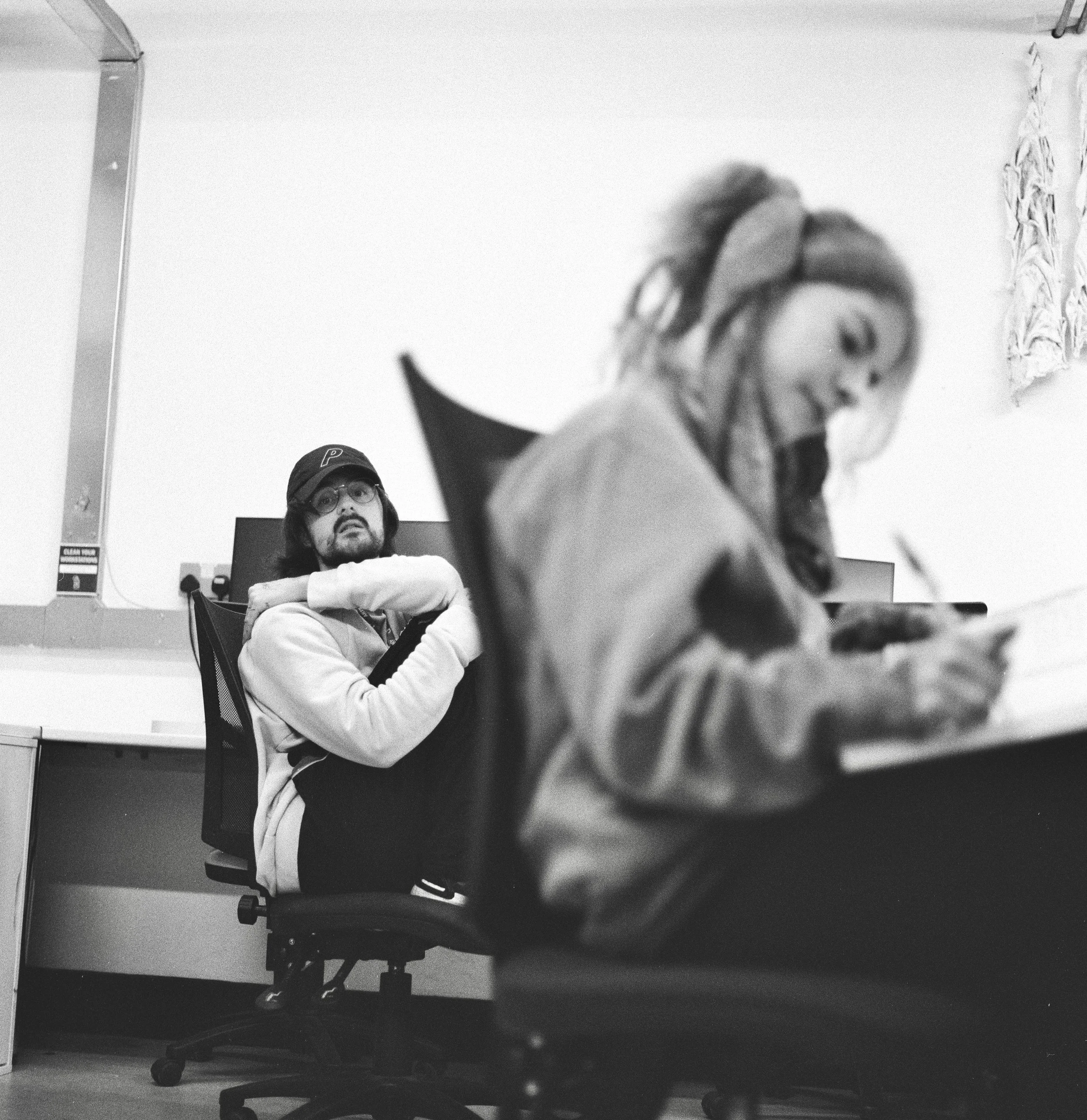 A black and white photo shows two men with long hair and glasses in a room with plain white walls. One man is sitting on a chair, looking forward, wearing a light-colored hoodie and a cap. The other man is standing, leaning over a desk or table, with