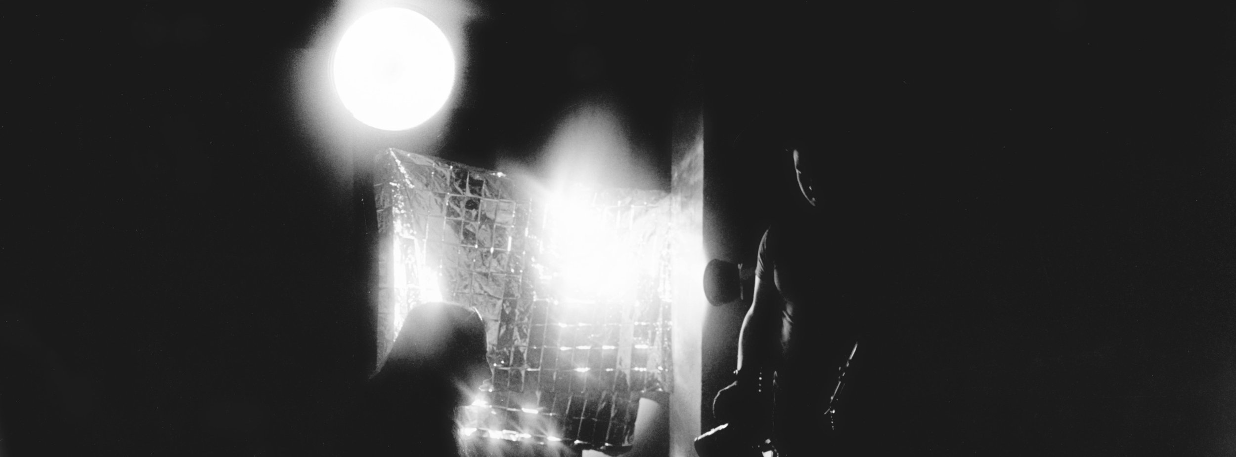 A dark scene with a bright light source illuminating a person in profile, showing their head and shoulder, seated on the right side, with reflective or metallic objects in front of them.