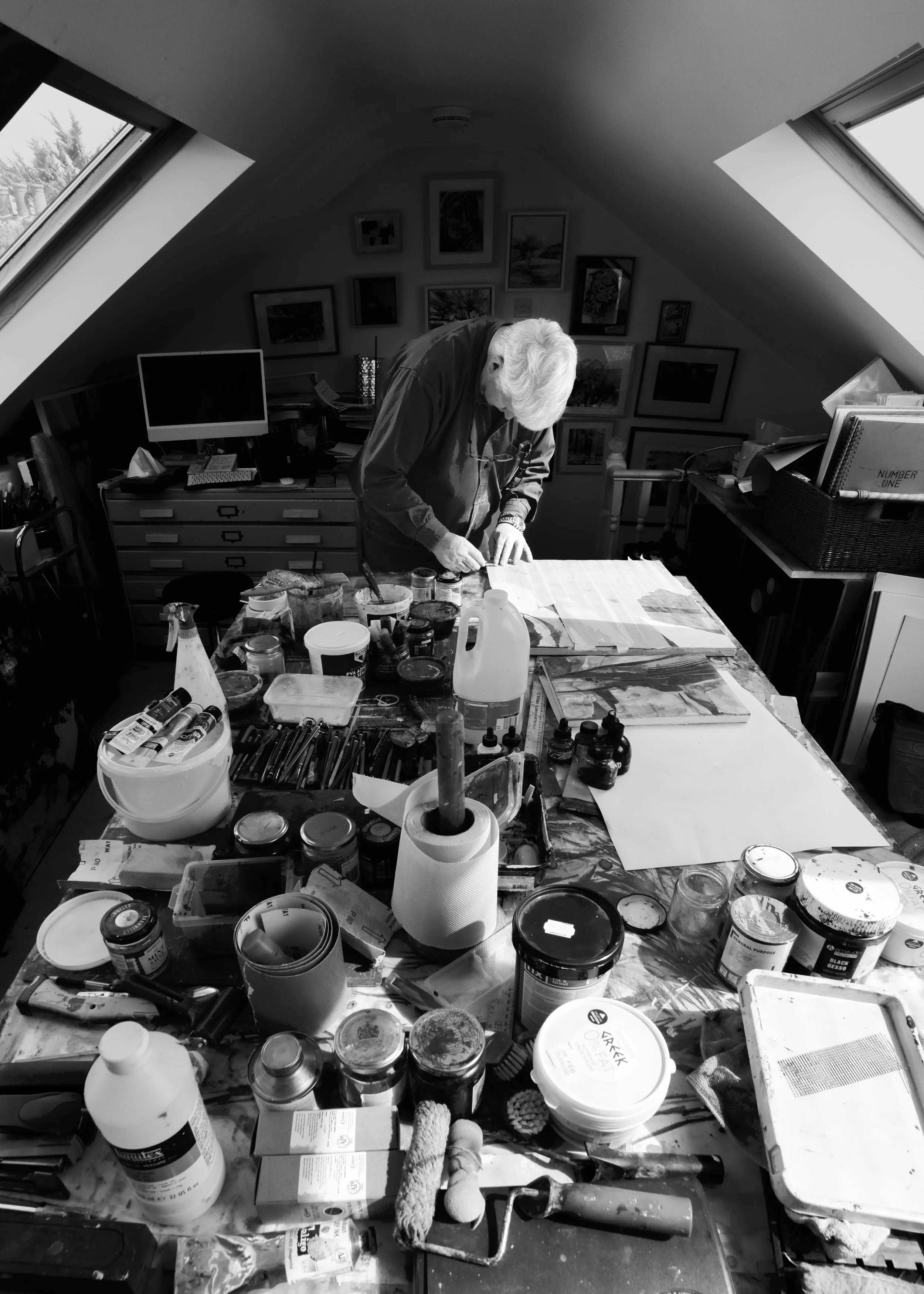 An elderly woman with white hair working at a cluttered table in a room with sloped ceilings and gallery walls. The table is filled with art supplies like paint jars, brushes, paper towels, and various containers.