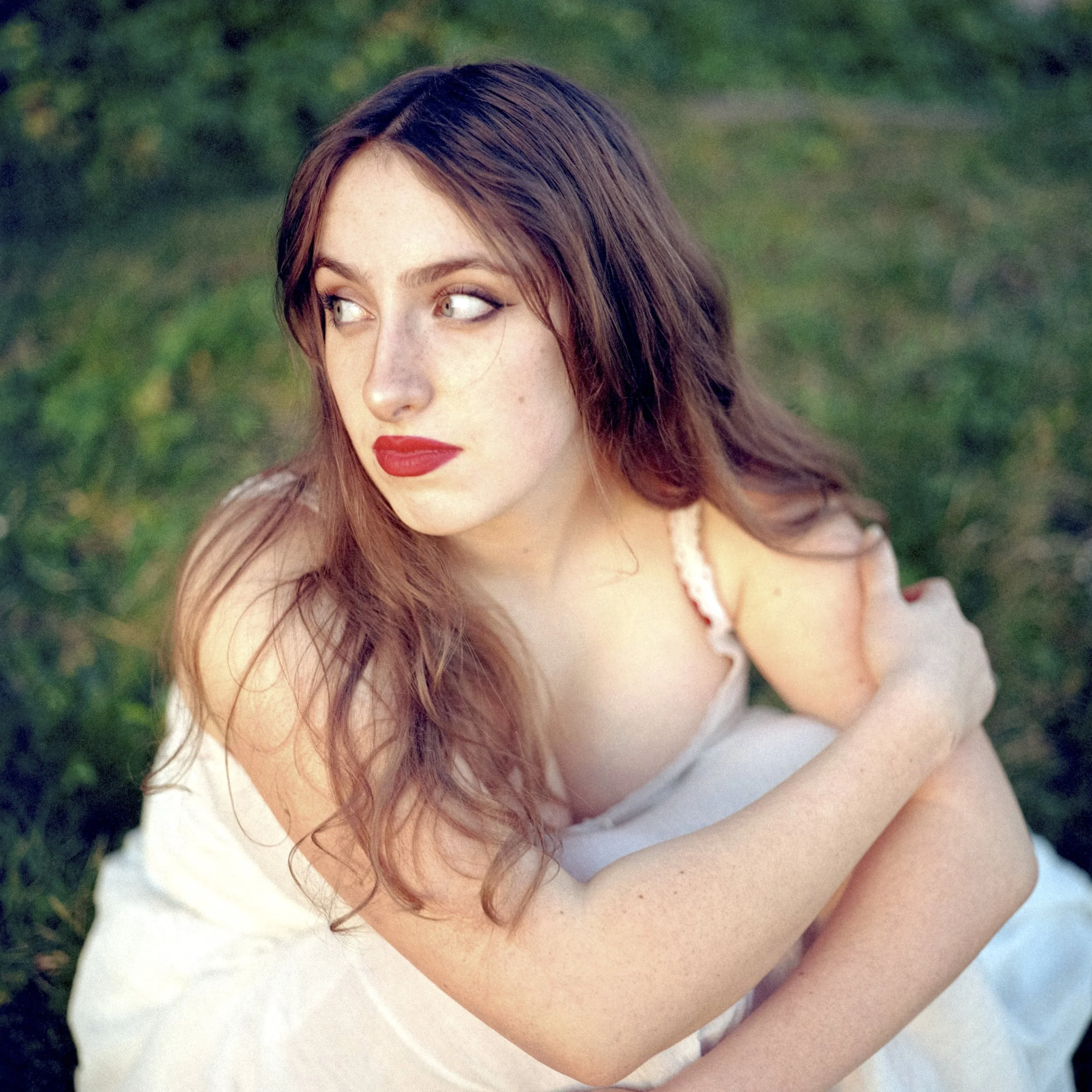 A close-up photo of a woman's face with red hair, wearing makeup including red lipstick, outdoors with green trees in the background.