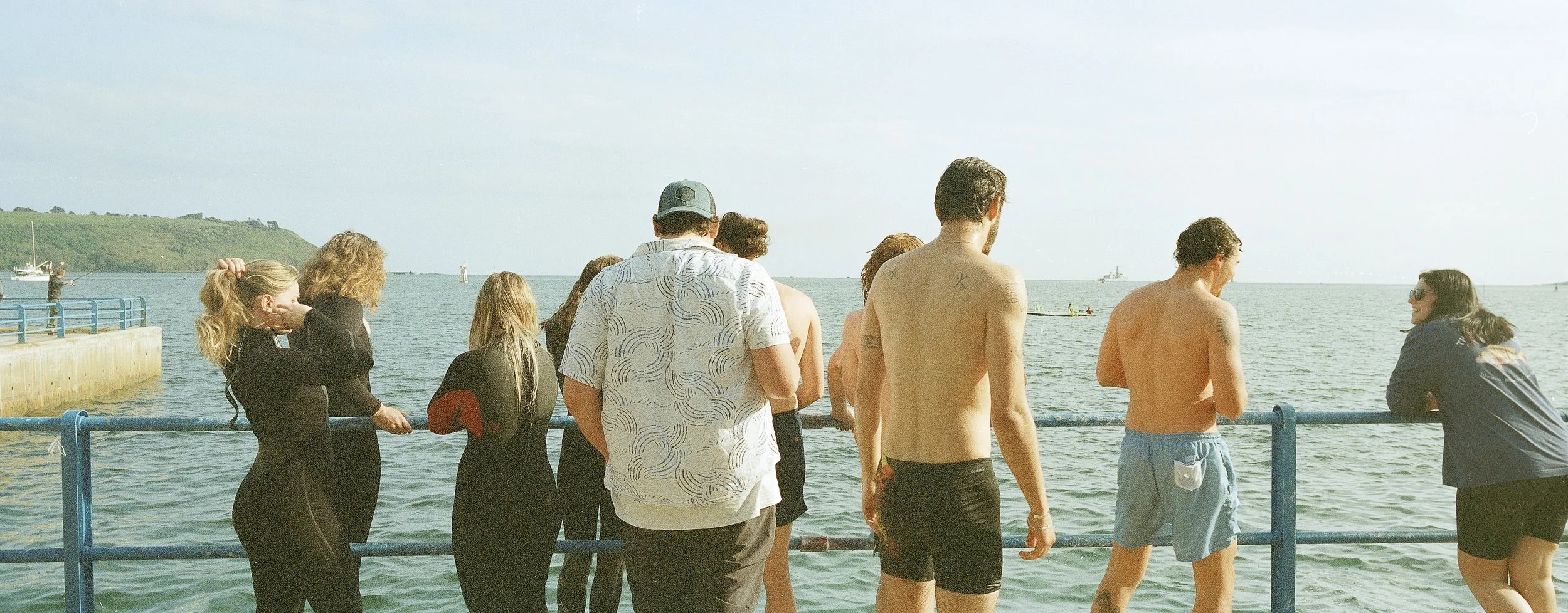 People standing on a pier near the water's edge during daytime, some wearing swimsuits or casual clothing, with boats and a distant green hillside in the background.