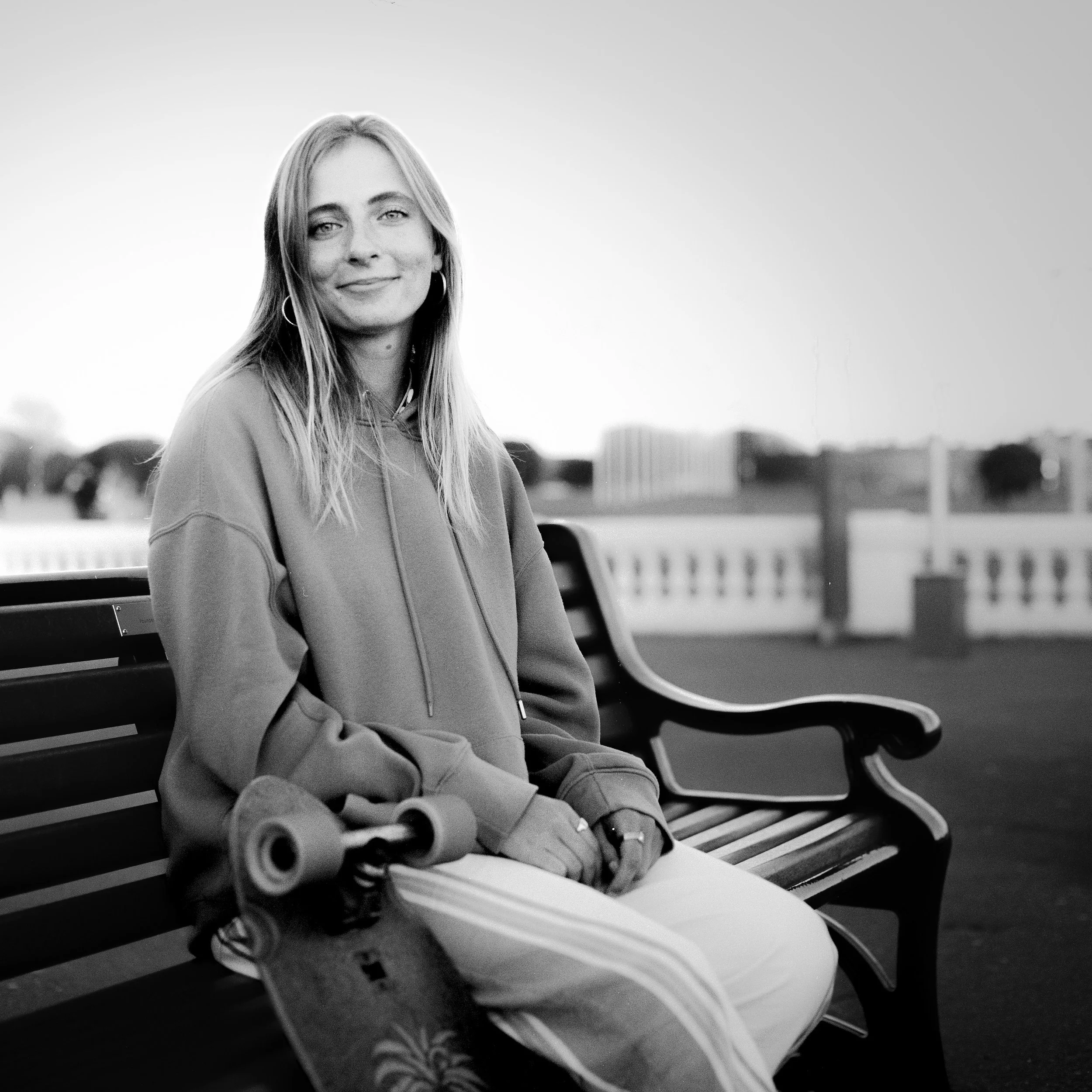 A black and white photo of a woman sitting on a bench with her arms crossed, smiling slightly, with a blurred background of buildings and a cloudy sky.