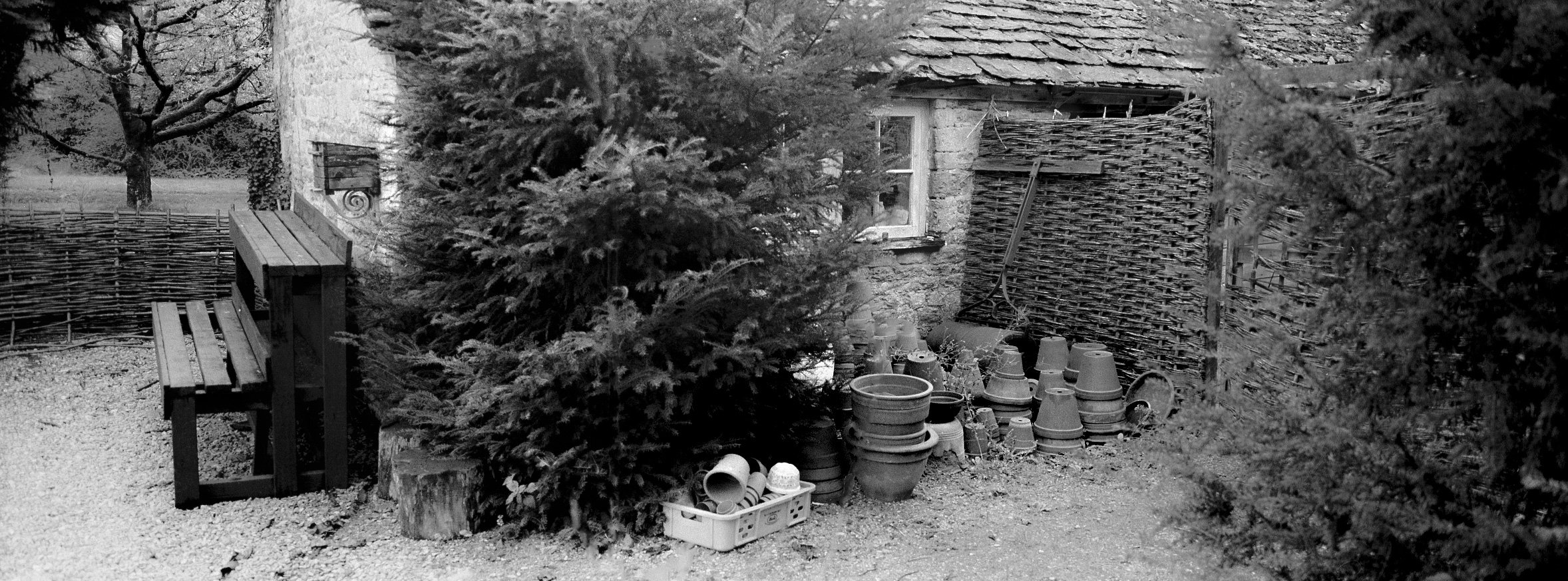 A rustic outdoor scene featuring a stone house with a small window, a wooden bench, and a pile of plant pots and gardening supplies near a small tree, surrounded by a woven wooden fence and trees.