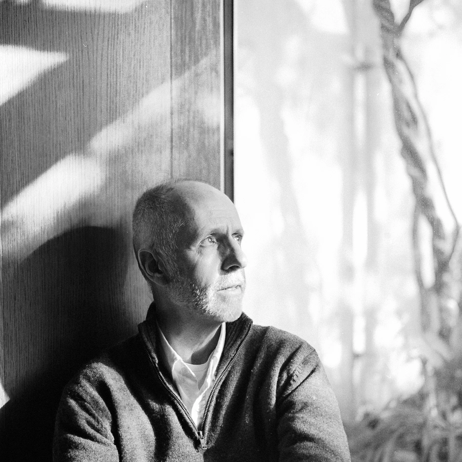 A black and white photo of a man with a beard and short hair, wearing a zip-up sweater over a collared shirt, looking to his right while sitting indoors. In the background, next to the man, there is a window or glass door, with part of the outside vi