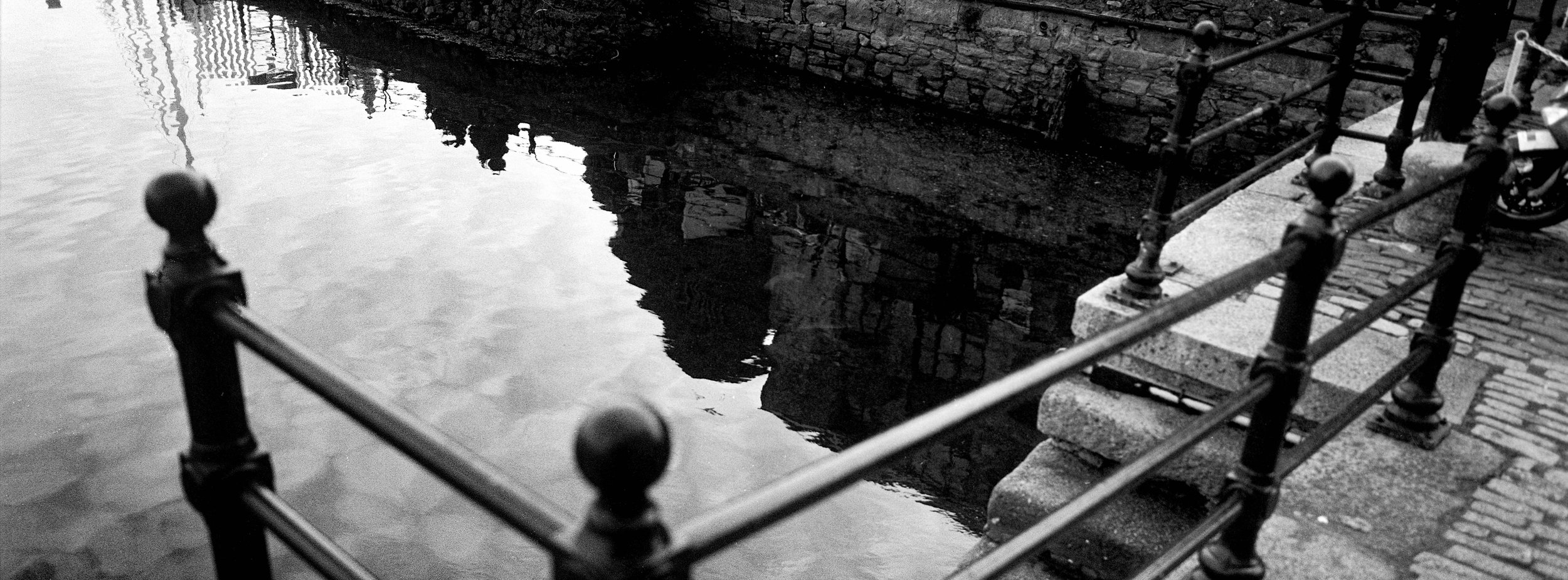 Black and white photo of a waterway with a stone embankment and a metal railing along the sidewalk.