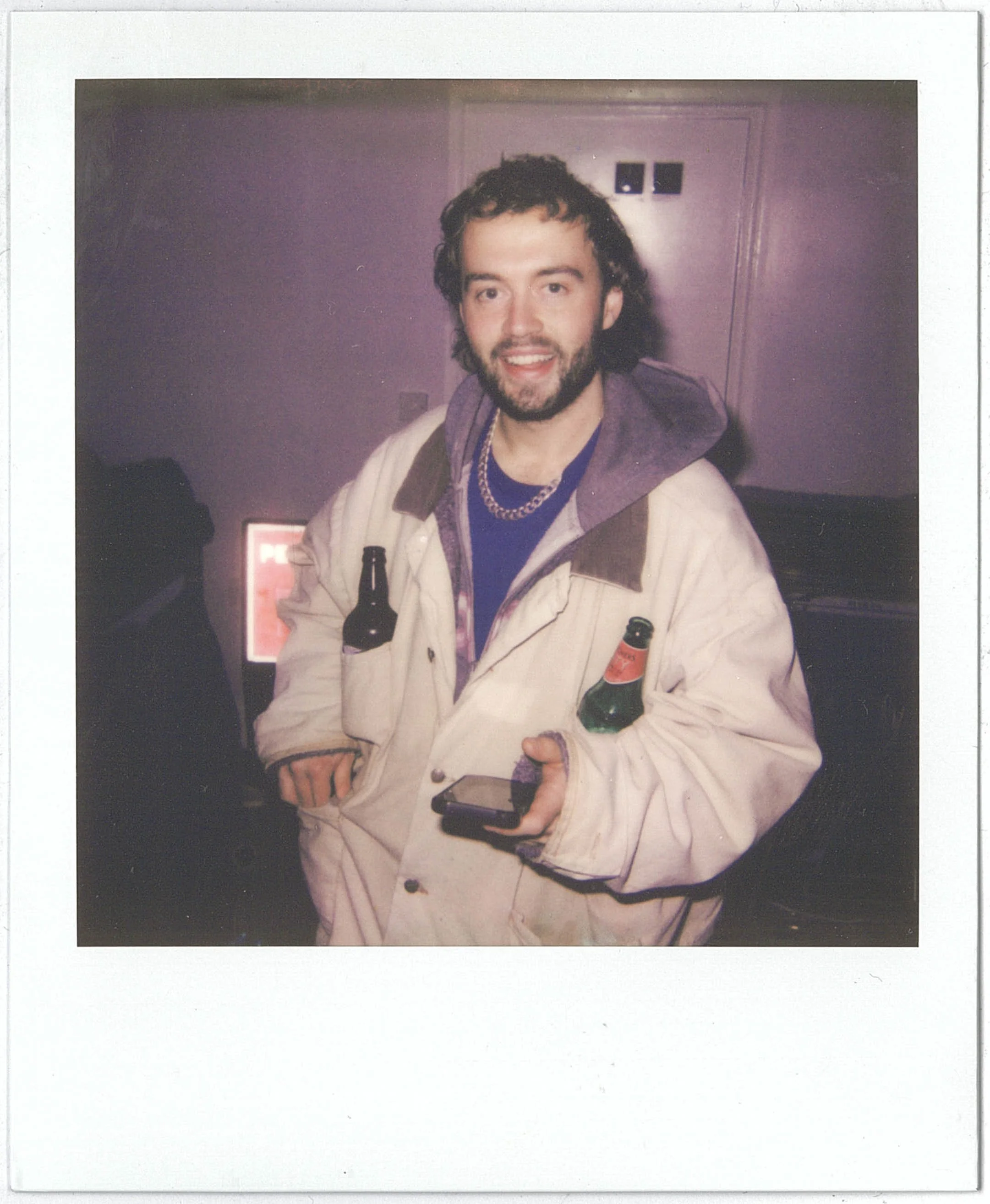 A Polaroid of a young man with curly hair, beard, and a wide smile holding a beer bottle and a cell phone in his hands, wearing a beige jacket and several necklaces, standing in a room with purple walls and a door in the background.