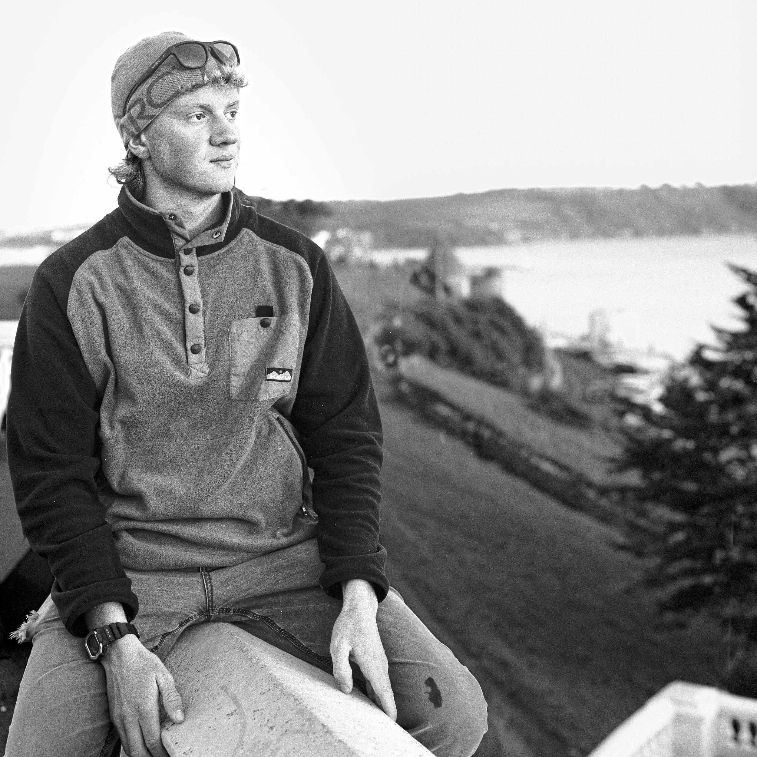 A person sitting on a large rock near a body of water, wearing a beanie, sunglasses on their head, a jacket, and a watch, gazing into the distance with a landscape in the background.