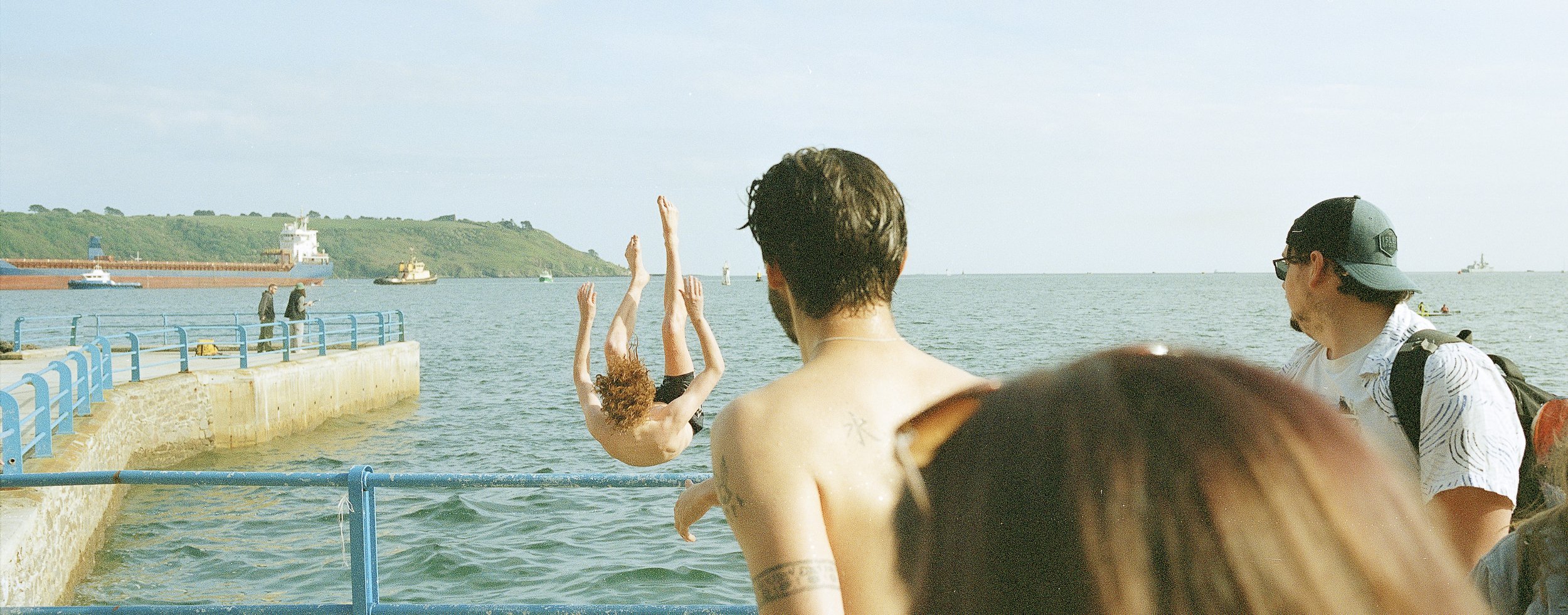A person diving into the water from a pier while others watch by the water's edge; boats and a green hill are visible in the background.