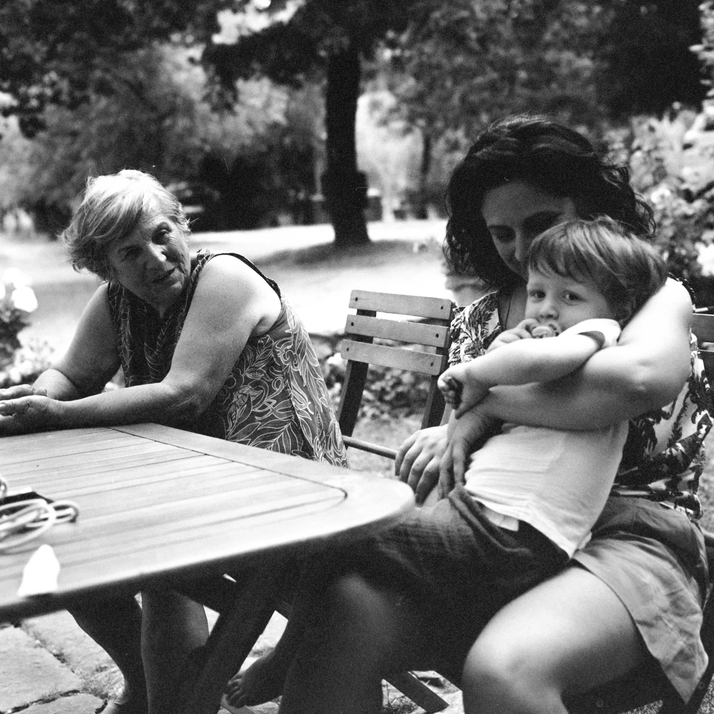A group of people sitting at a table outdoors, including an elderly woman with light hair, a girl with curly hair, and a young boy, in a park or garden setting.