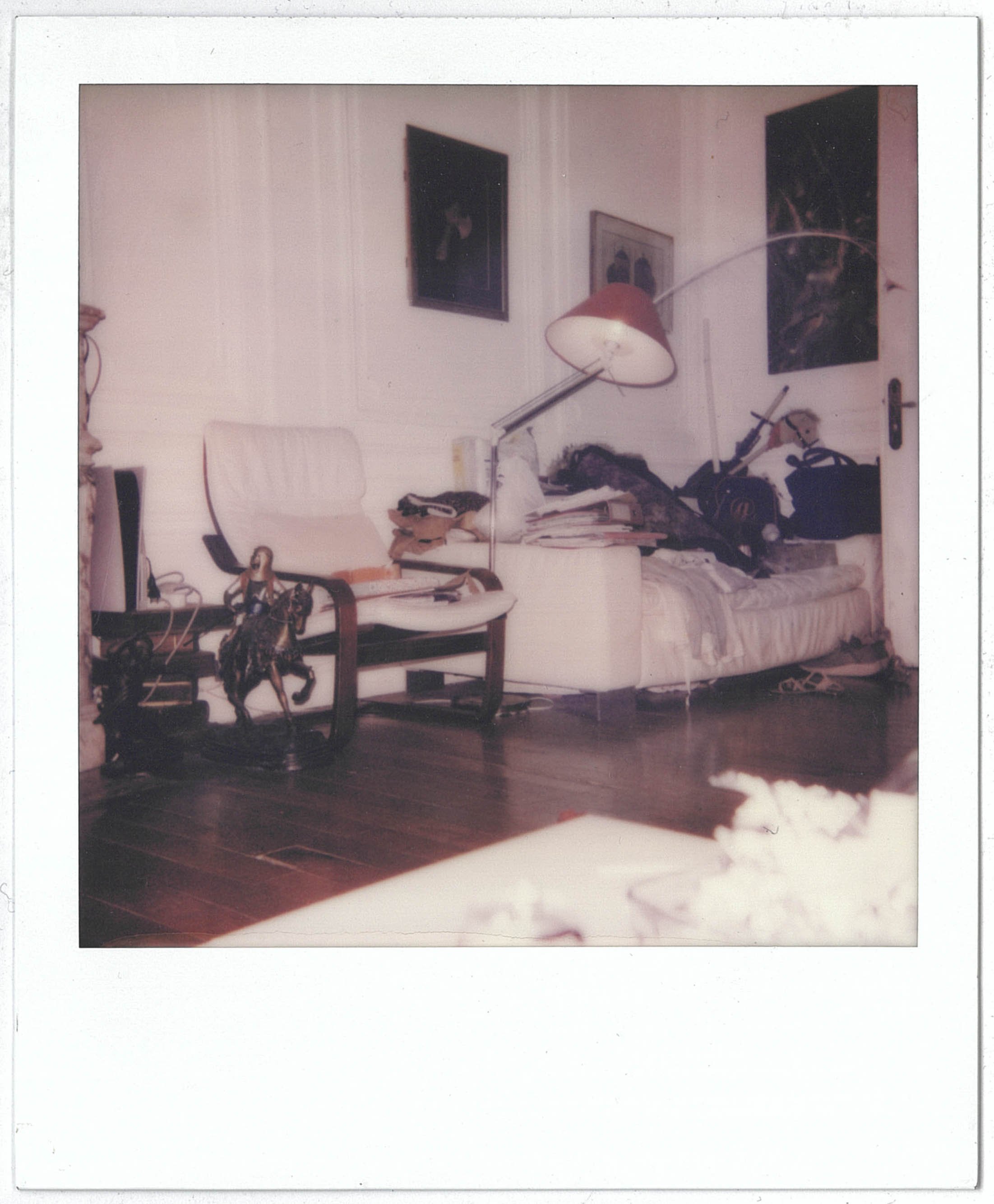 A Polaroid of a cluttered bedroom with a bed, chairs, and various objects, including clothes, bags, and a lamp, with wallpapered walls decorated with framed pictures and paintings.