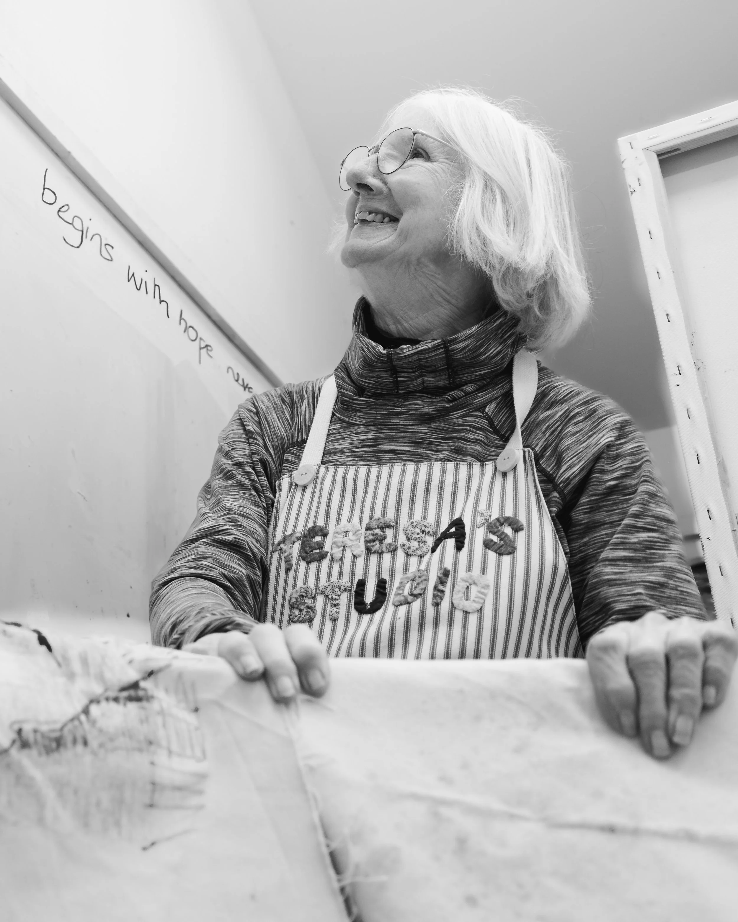 Smiling elderly woman with glasses wearing an apron, preparing baked goods in a kitchen.