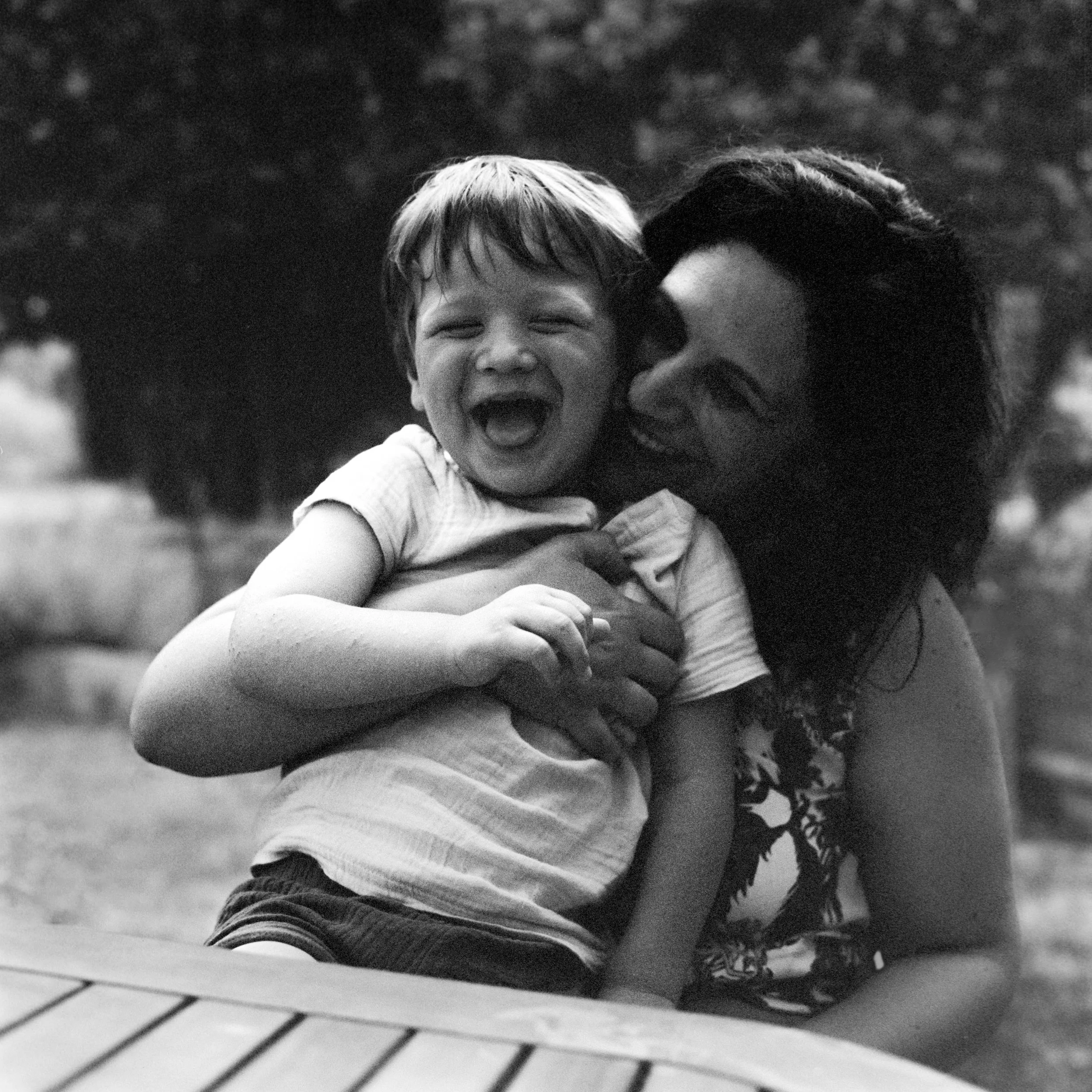 A black and white photo of a smiling child being embraced by a woman, with trees in the background and a tiled surface in the foreground.