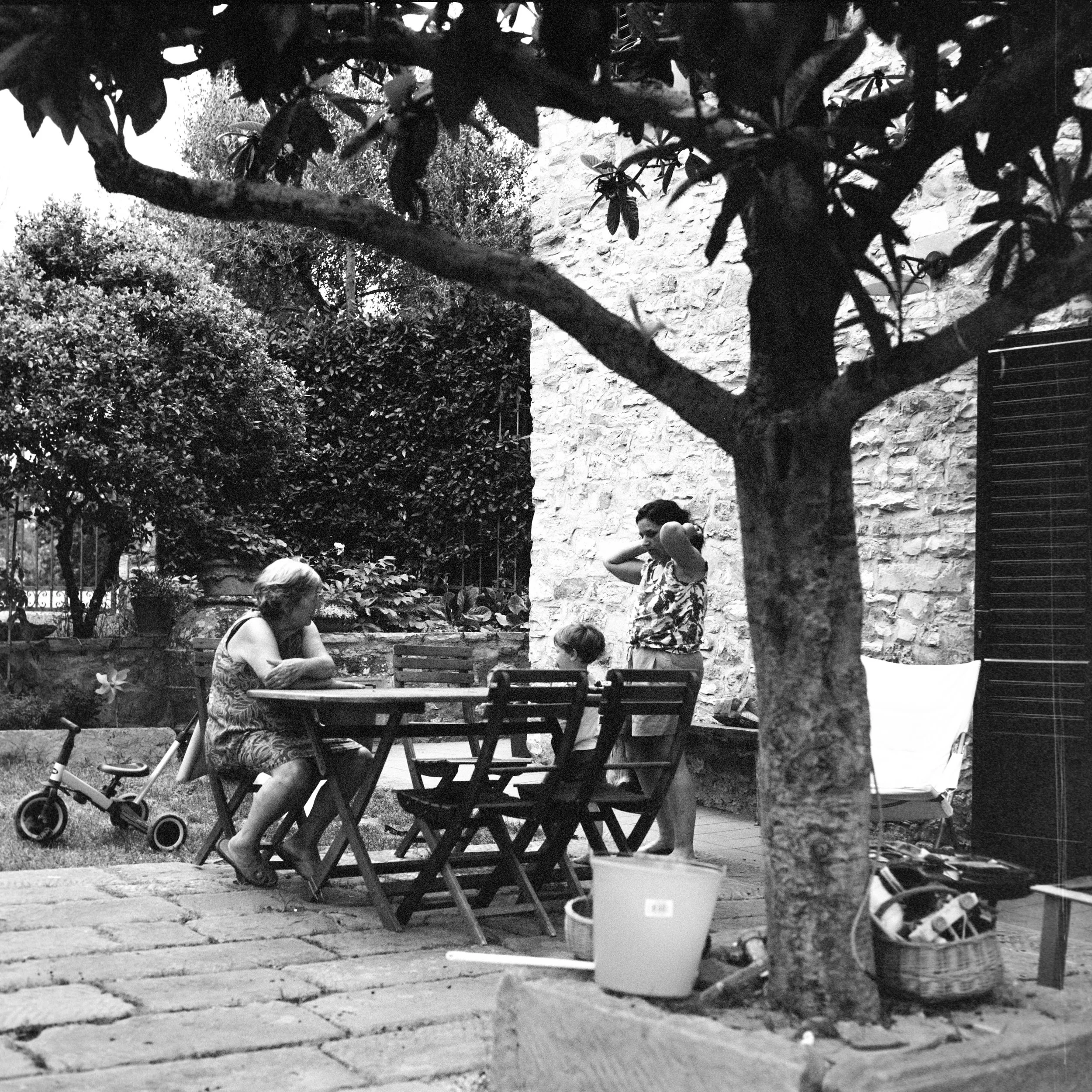 Children playing outdoors next to a tree and a stone wall in a garden, with a woman sitting at a picnic table.
