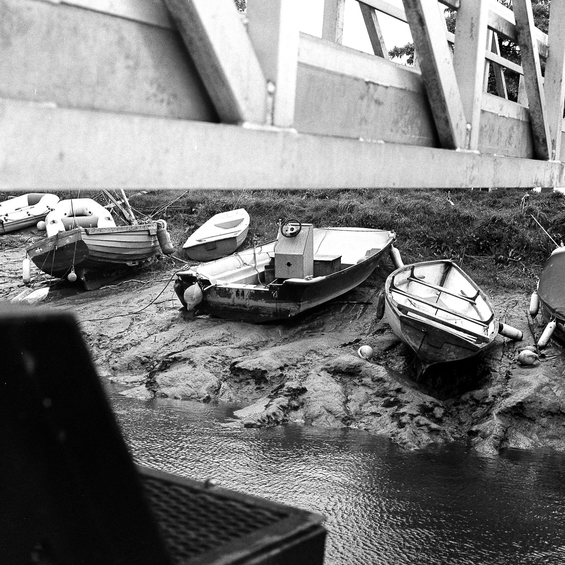 Various boats are docked on a muddy shoreline near water, with a large wooden structure, possibly a dock or pier, extending over the scene. Trees are visible in the background.