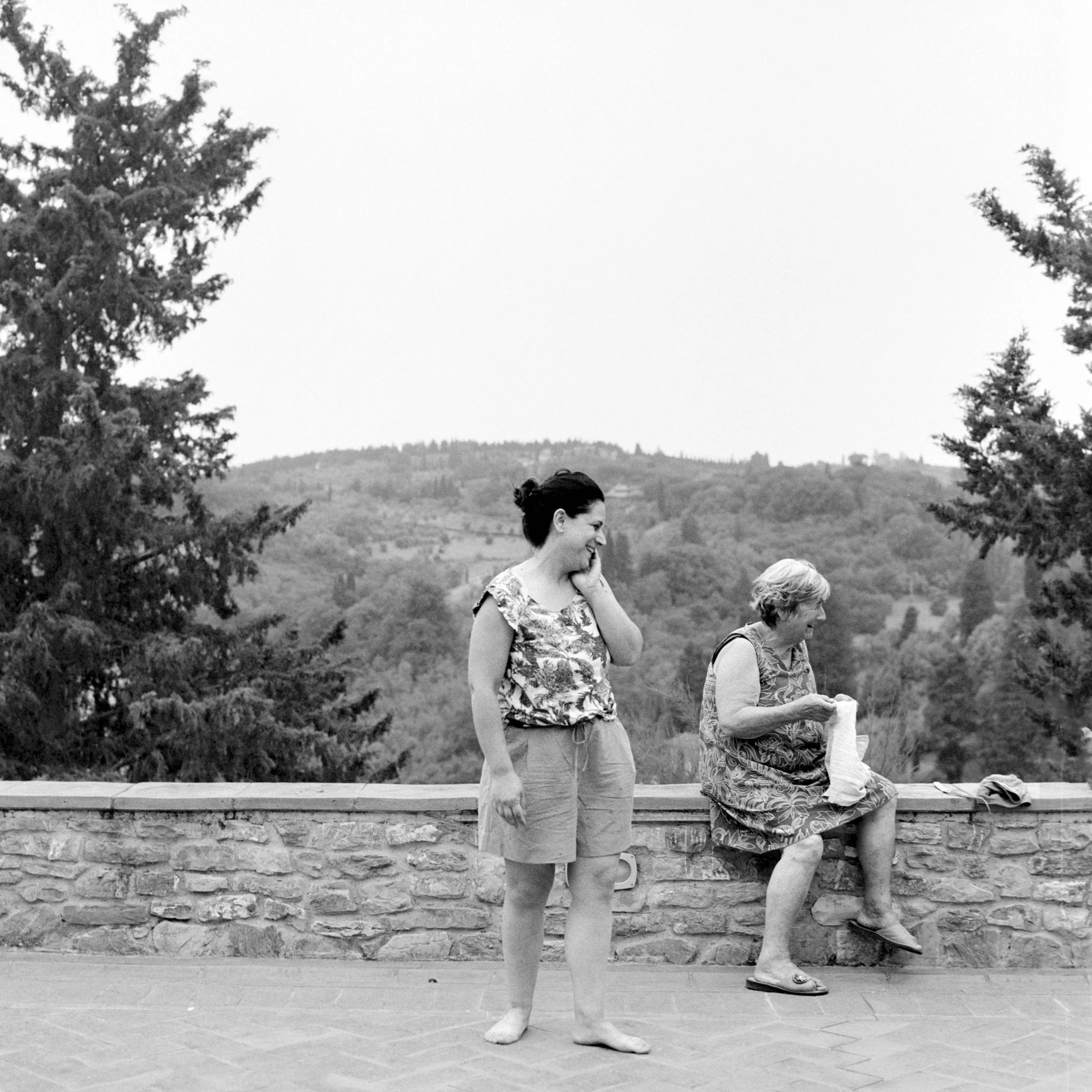 A black and white photo of two women walking along a stone wall, with trees and a distant hill in the background. The women are smiling and appear to be enjoying themselves.