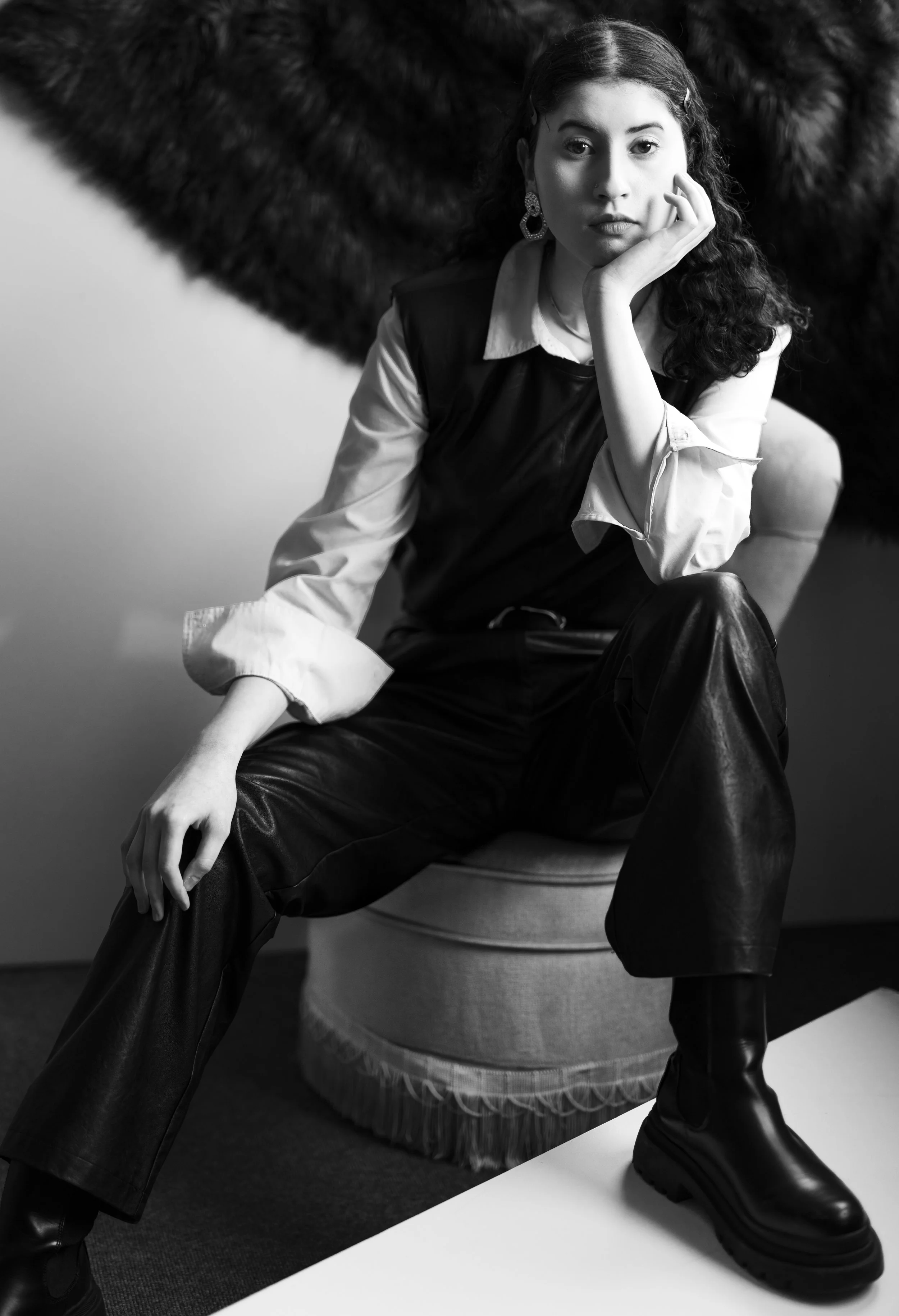 A woman sitting on a fuzzy chair, wearing a leather outfit and a white shirt with rolled-up sleeves, black leather pants, and black boots, with her chin resting on her hand and a serious expression.