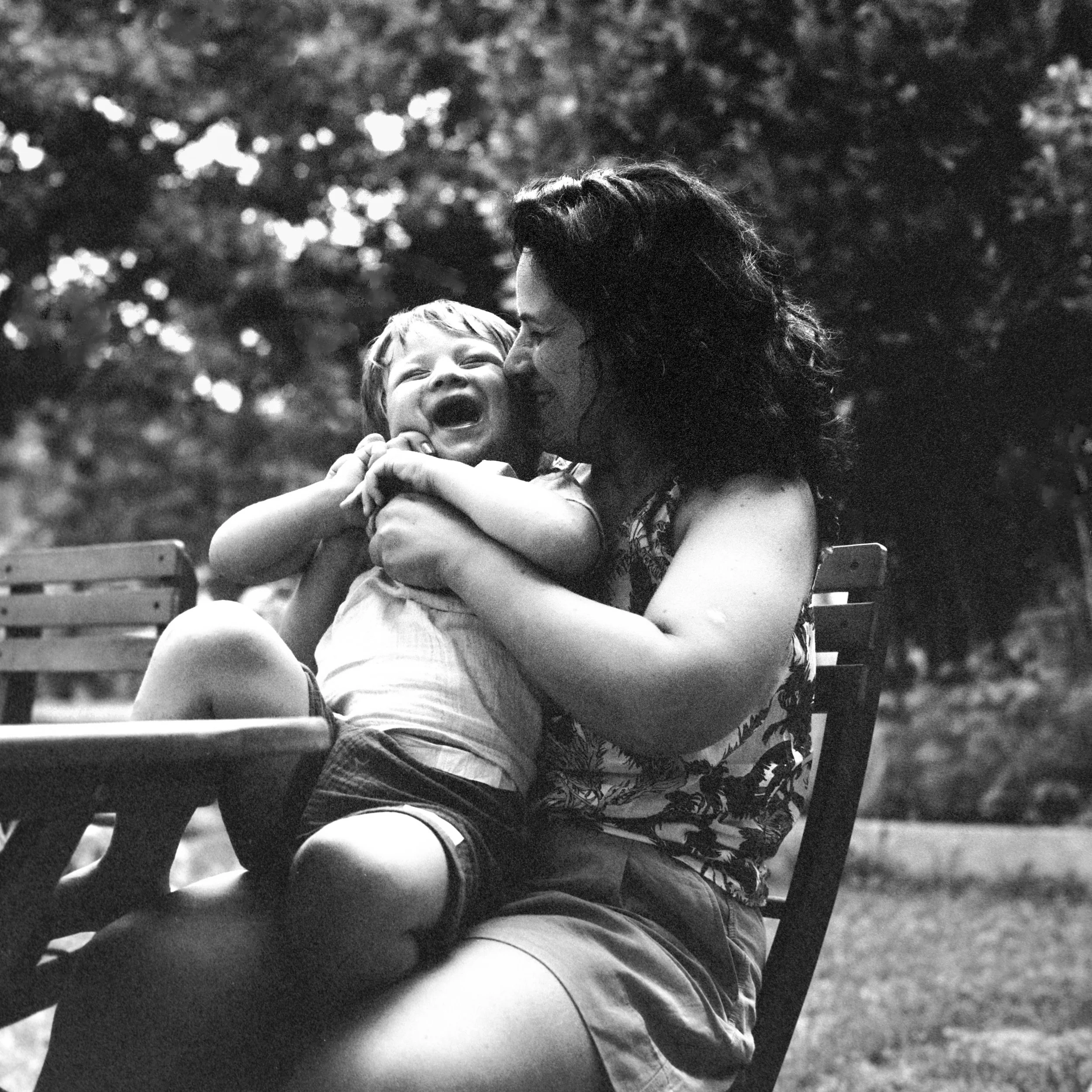A black-and-white photograph of a woman and a young child sharing a tender moment outdoors. The woman is kissing the child's cheek as the child laughs joyfully, showing teeth. They are sitting on a park bench with trees blurred in the background.