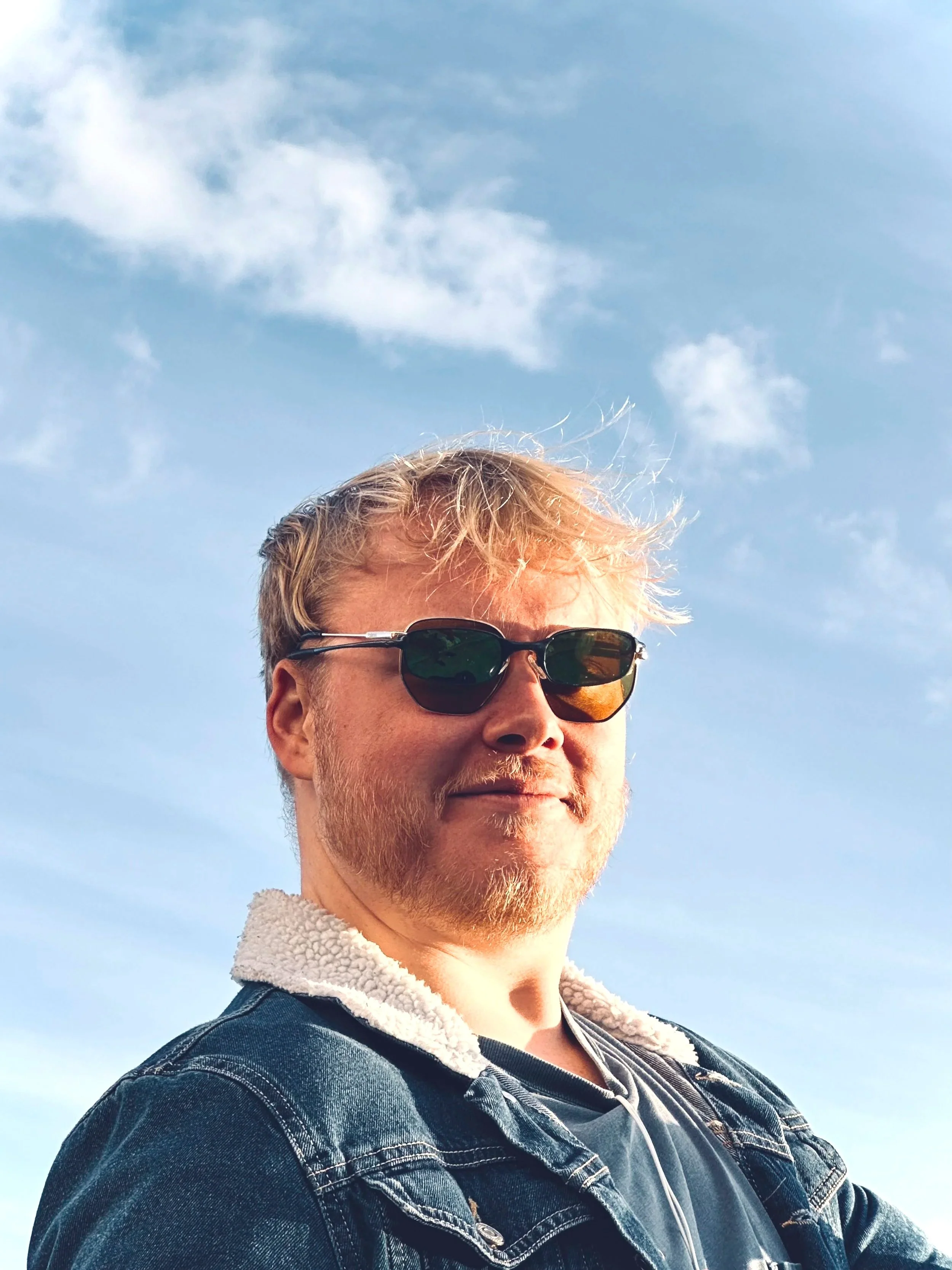 A person with light-colored hair wearing sunglasses, a denim jacket, and a white fleece collar, smiling against a blue sky with scattered clouds.