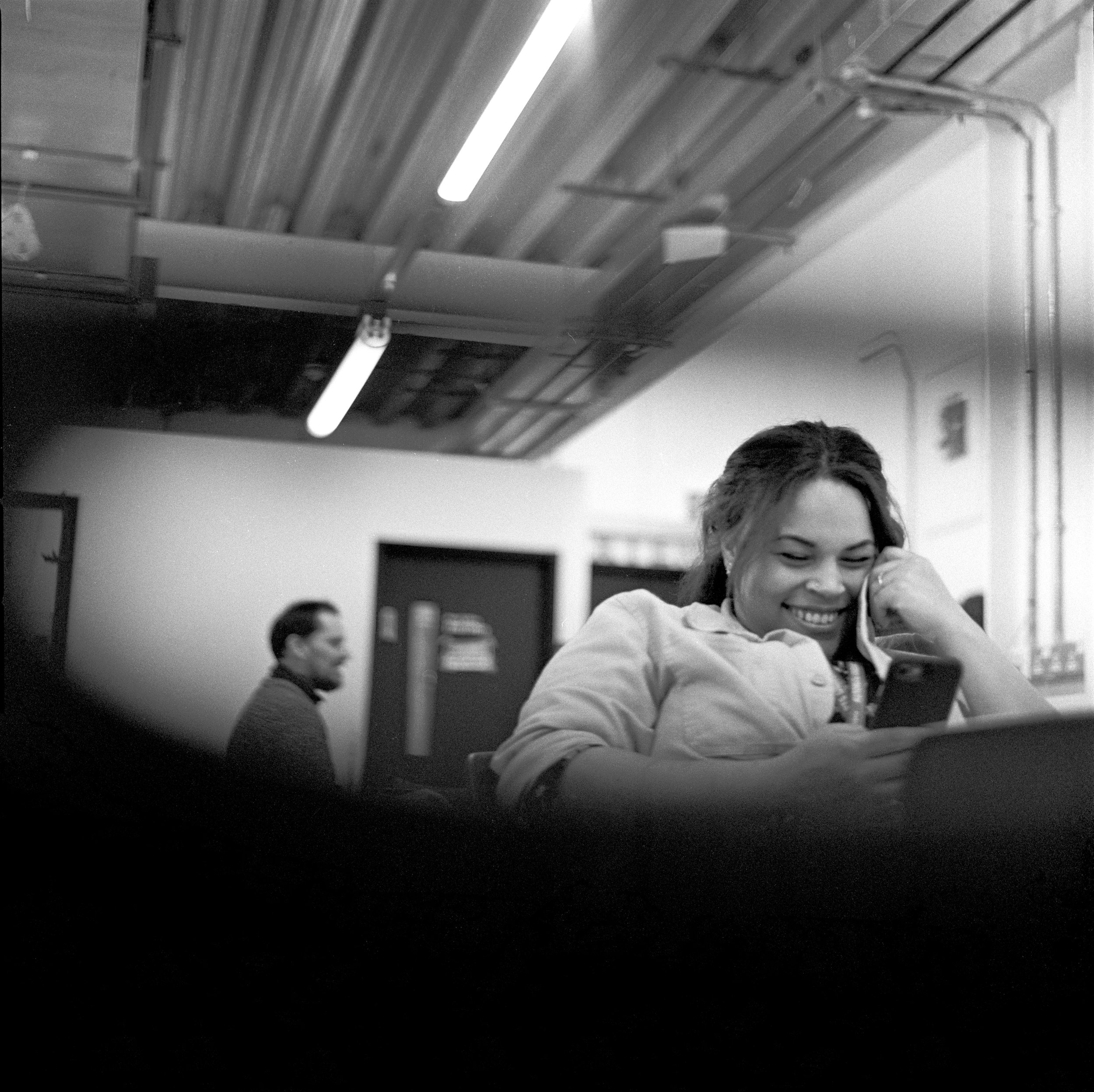 Black and white photo of people sitting in a room with a ceiling having fluorescent lights and visible pipes. One woman smiling and talking on the phone, another person in the background. The focus is on the woman in the foreground.