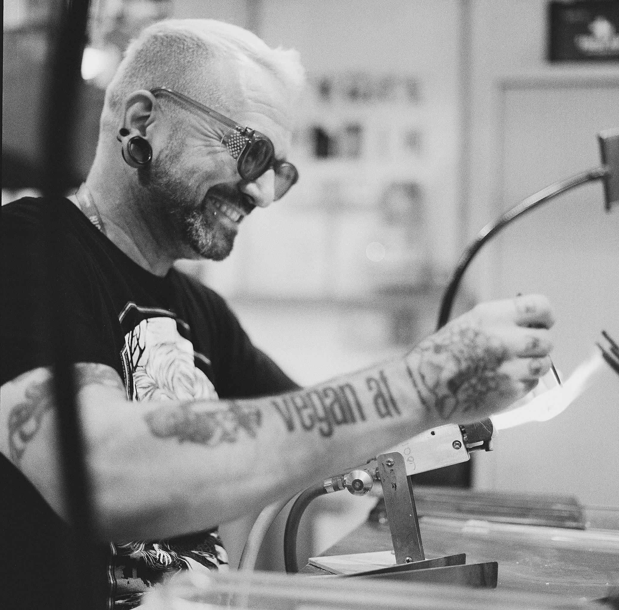 A person with short hair and stretched earlobes wearing glasses is seated at a table in what appears to be a public space or cafeteria. The person is smiling and looking down at a piece of paper in front of them. The image is in black and white.
