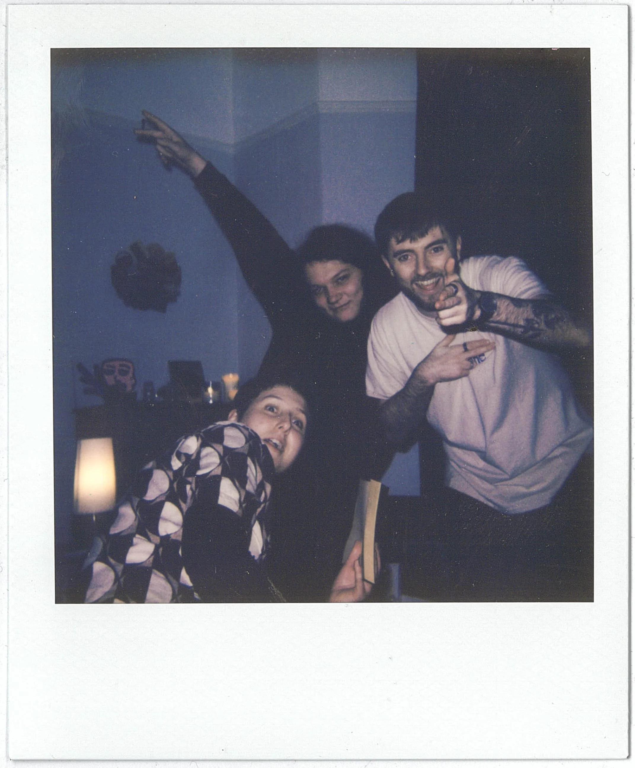 A Polaroid of two young men smiling and posing for the camera. One is pointing at the camera with his finger. They are in a dimly lit room with blue walls and some decorations in the background.