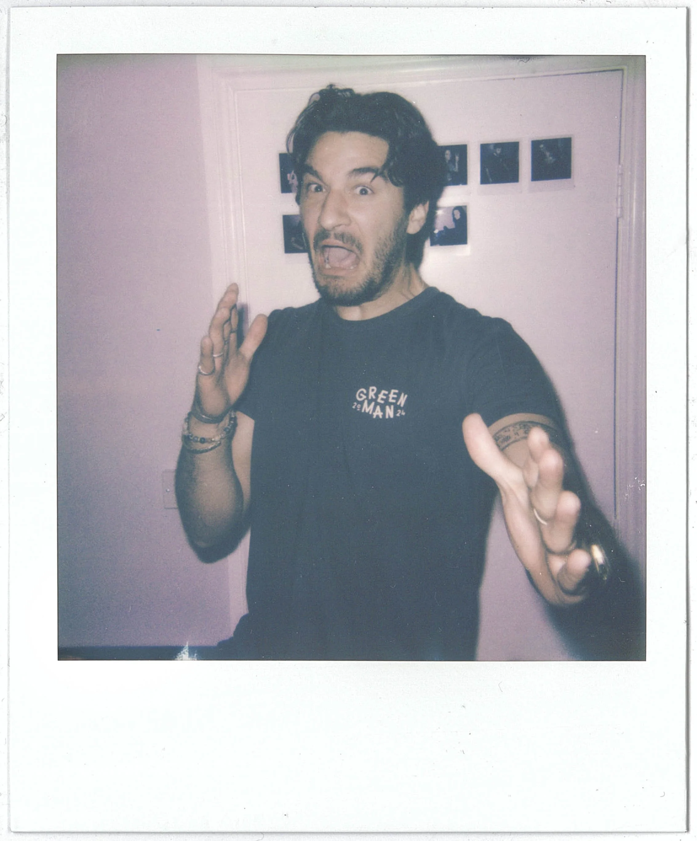 A person with dark, wavy hair and facial hair is making a surprised or excited facial expression in front of a pink wall with small framed photos. The individual is wearing a black T-shirt with white text and has multiple bracelets on their wrist.