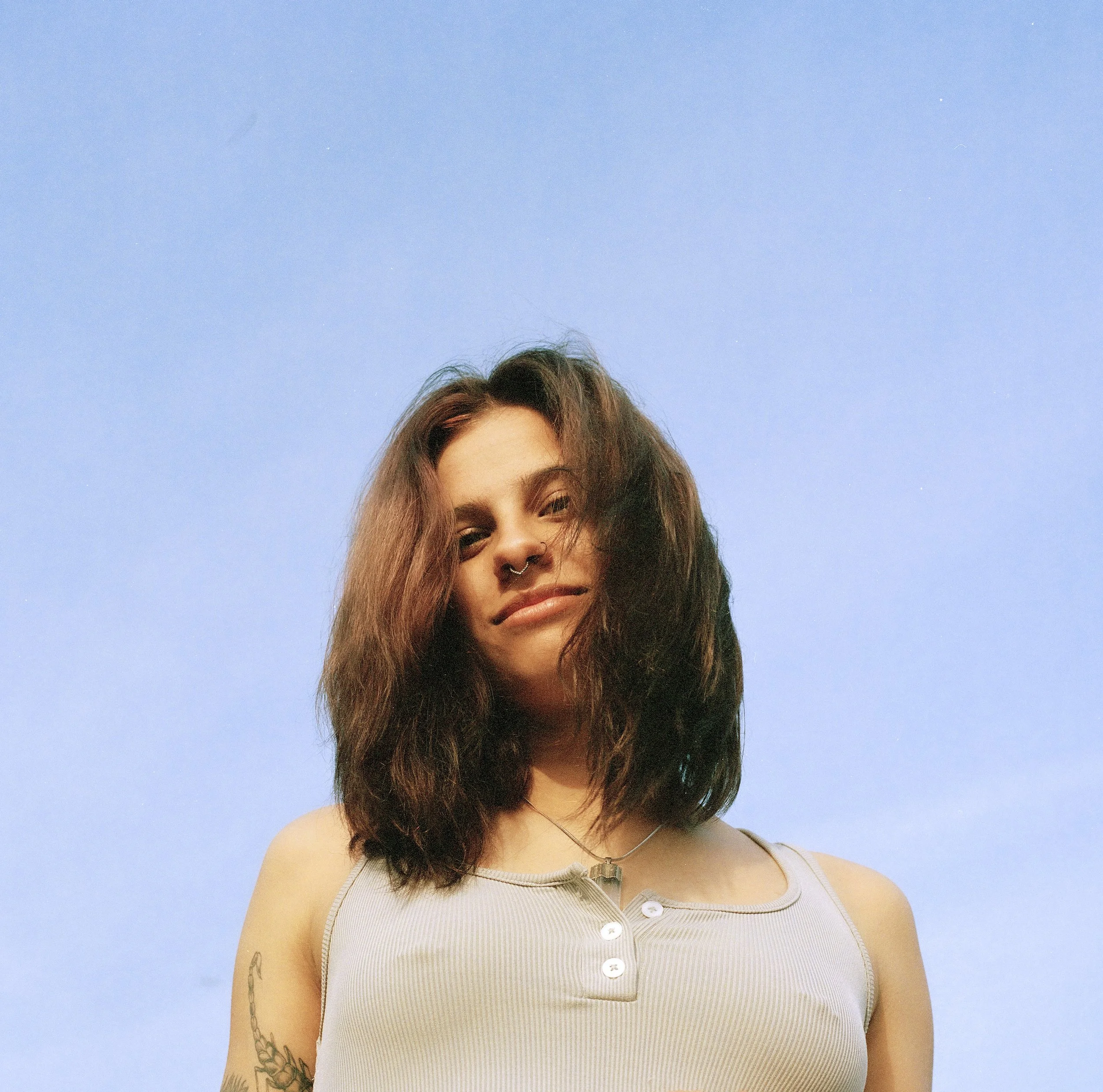 A woman with wavy brown hair, a septum piercing, and tattoos on her arm, wearing a light-colored tank top with buttons, against a blue sky background.