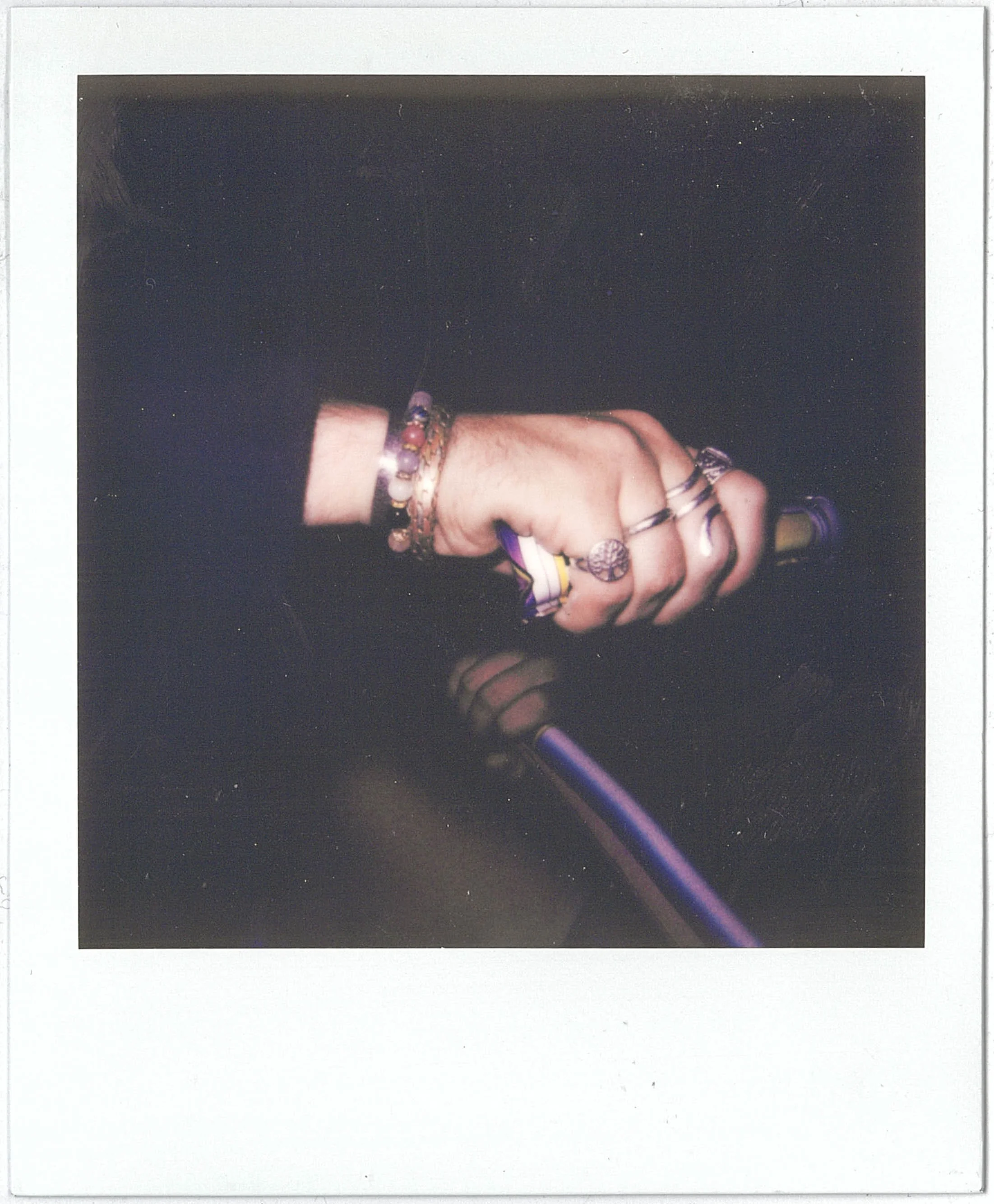 A Polaroid of a photograph of a hand holding a smartphone, wearing multiple bracelets and rings, with a dark background.