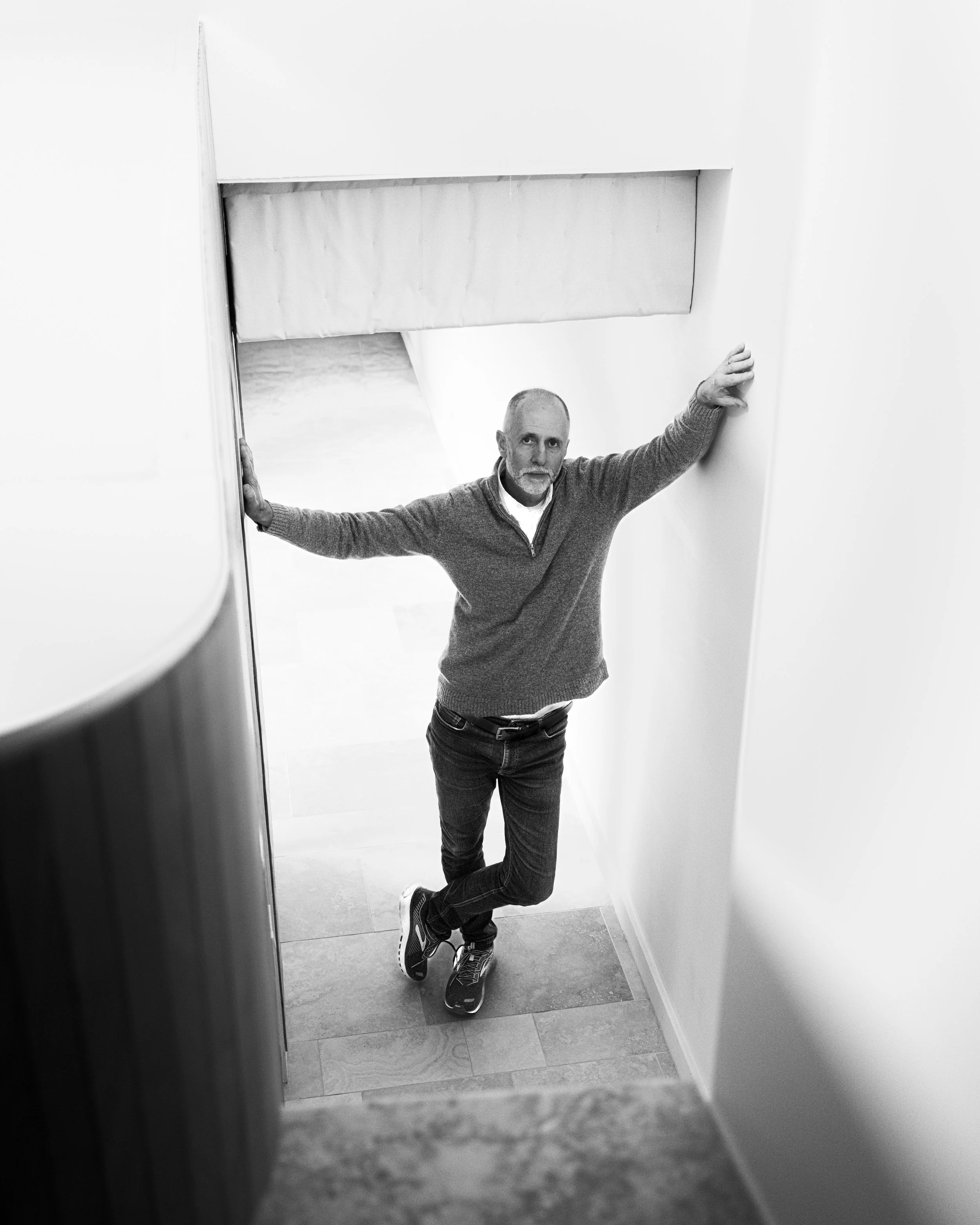A man with a beard and short hair in a gray sweater, black pants, and sneakers stands on a skateboard, leaning against a wall and holding onto the door frame in a narrow hallway.