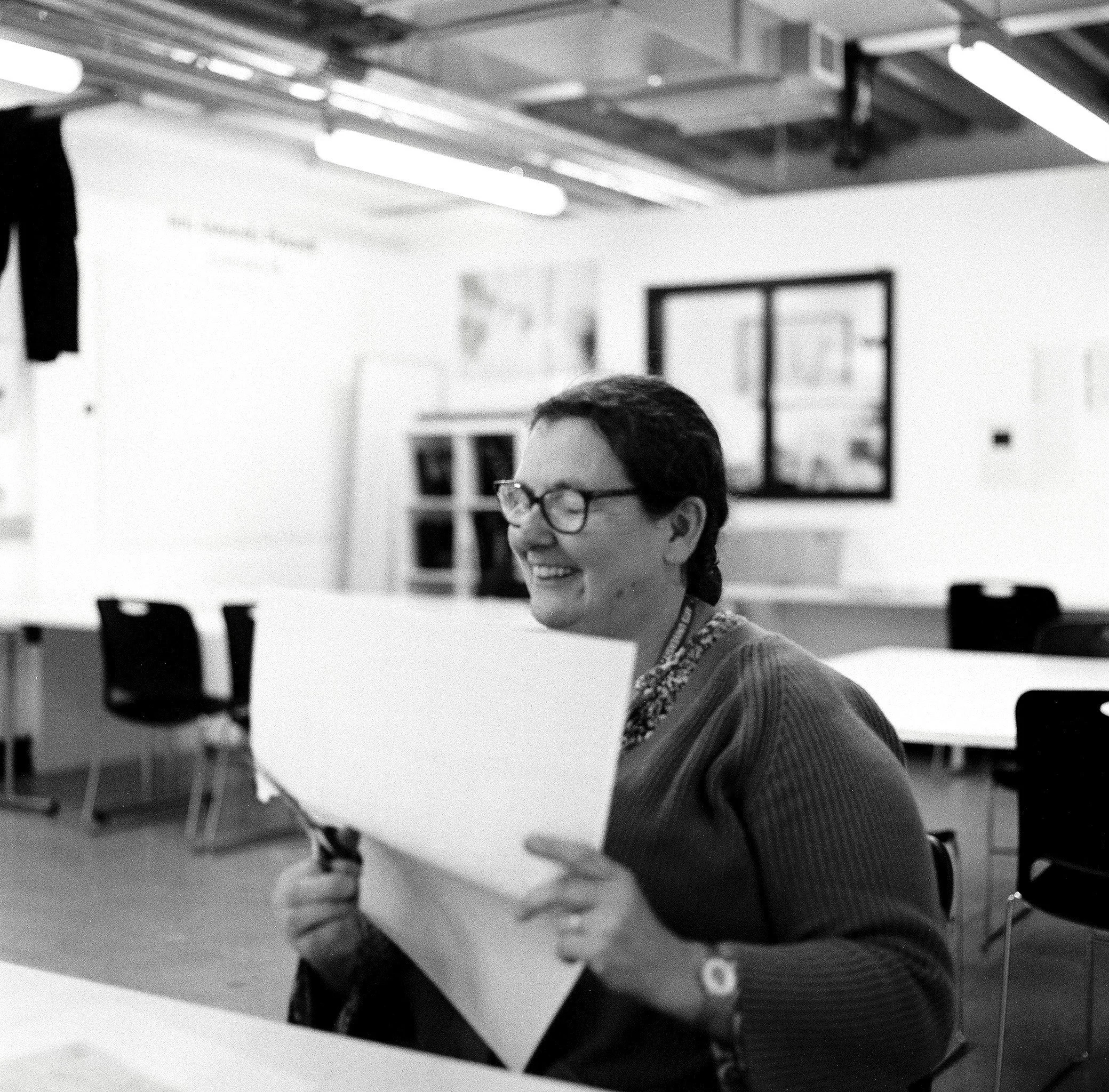 Black and white photo of a woman with glasses and a tattoo on her neck, smiling and sitting in a room with a basketball hoop on the wall, a framed picture, and chairs with a table in the background.