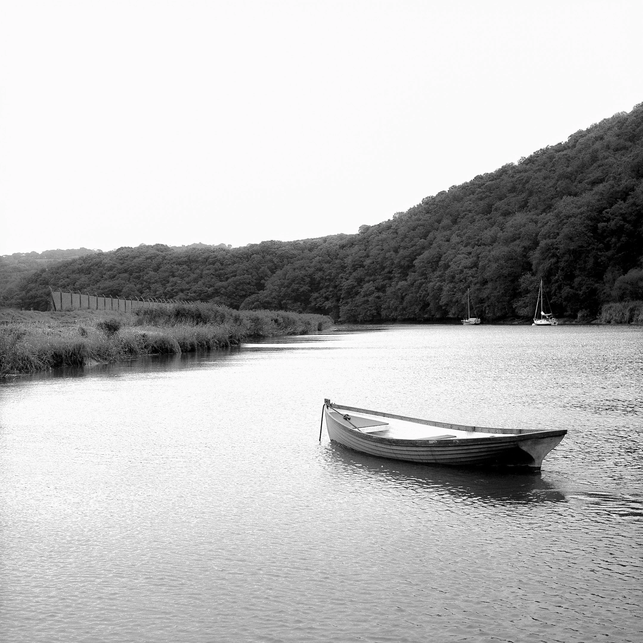 Black and white photo of a river with three sailboats anchored near the shore, surrounded by forested hills.