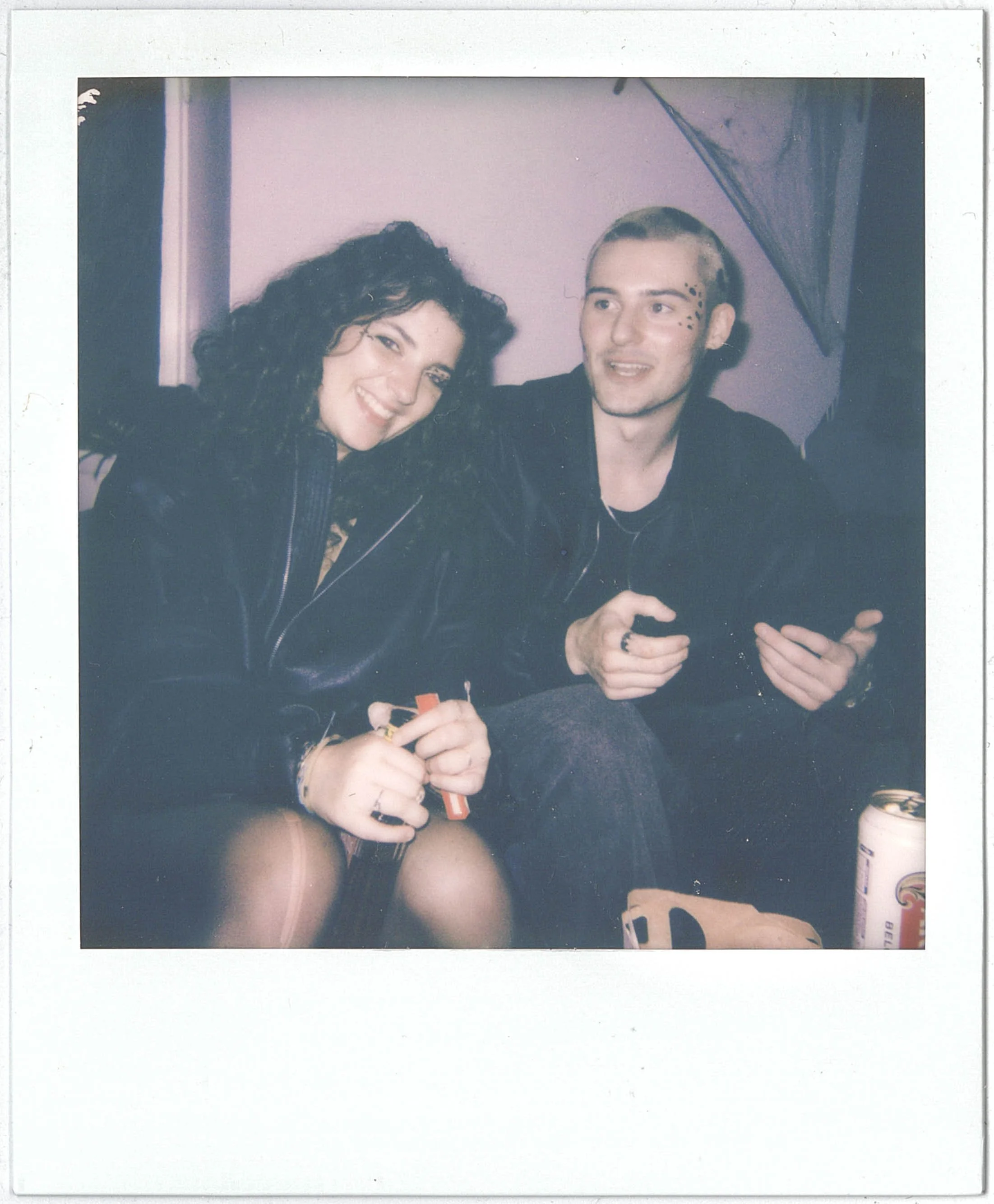 A Polaroid photograph of a smiling woman with curly hair and a man with short hair and facial ink designs, sitting together against a purple wall, with a rainbow-colored umbrella in the background.
