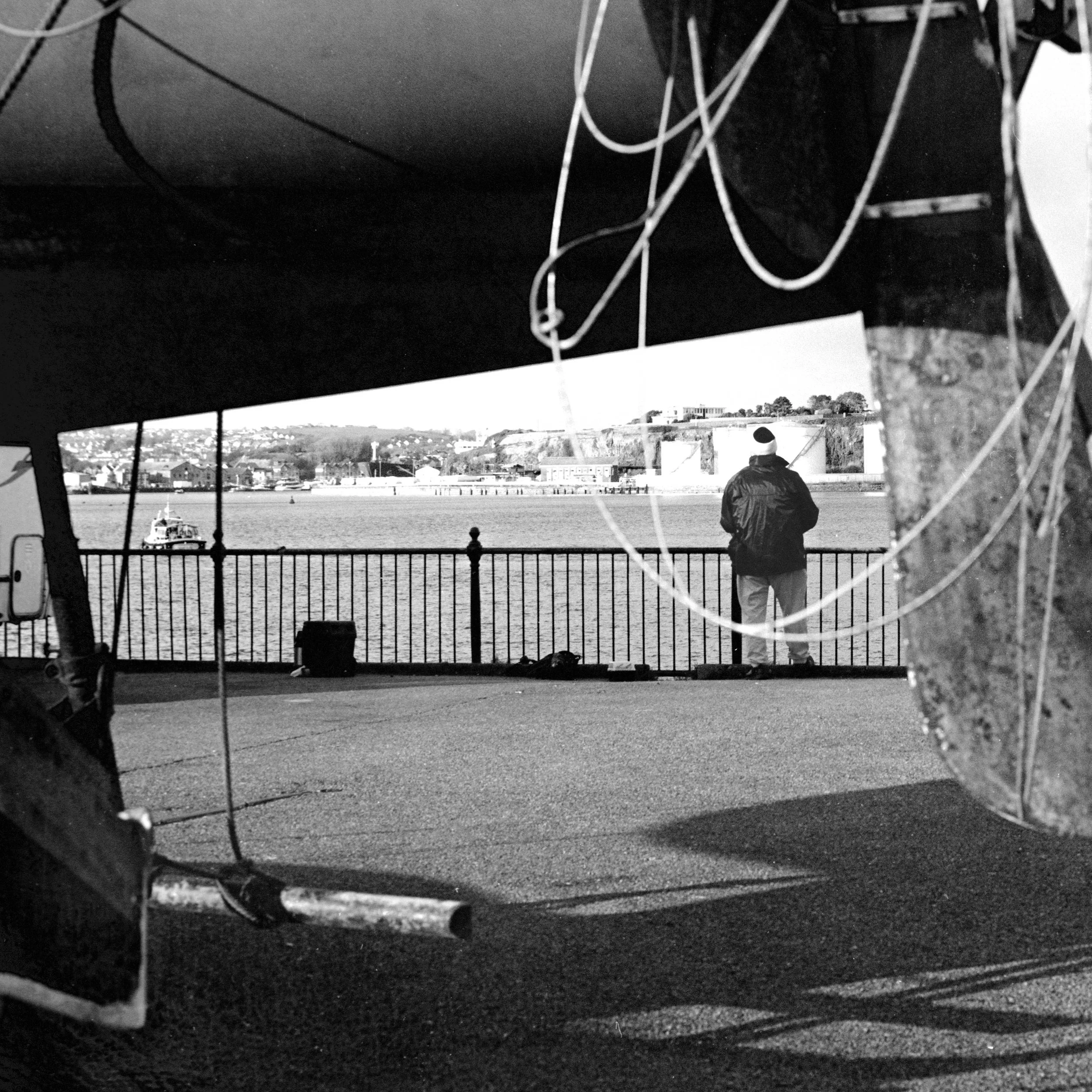 Black and white photo taken from under a boat, showing a person near the water's edge with boats and buildings in the background.