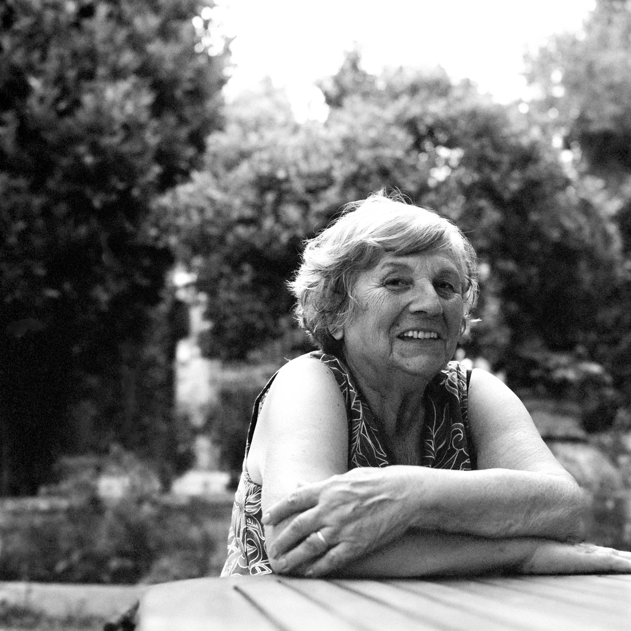 Black and white photo showing the top of a person's head, with short hair, in the foreground. The person is in front of a blurred background of trees and sky. The photo appears to be taken outdoors.