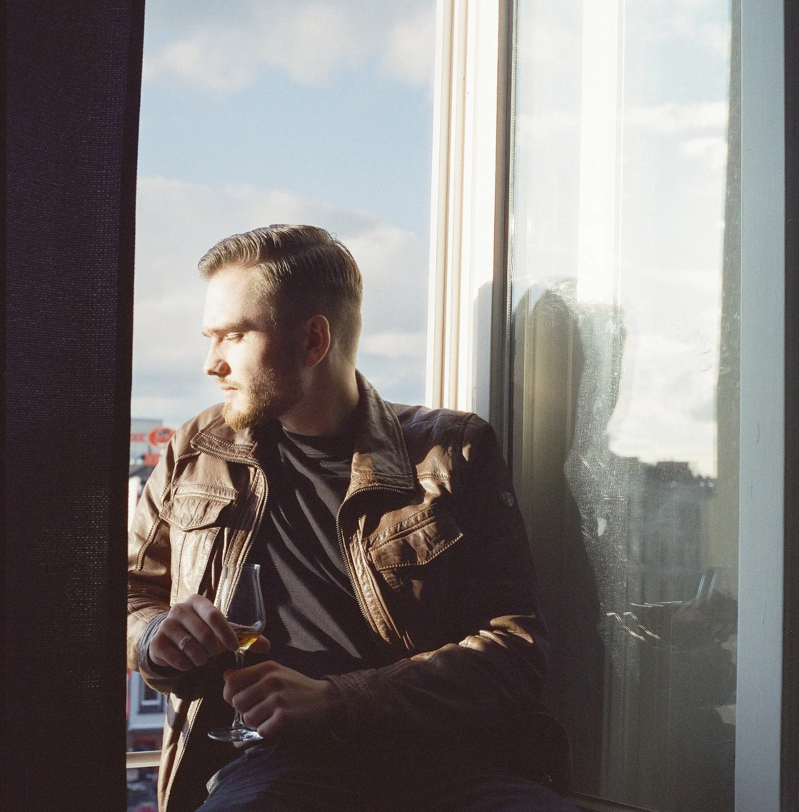 Young man with a beard and slicked-back hair sitting by a window, holding a glass of wine, looking outside on a sunny day.