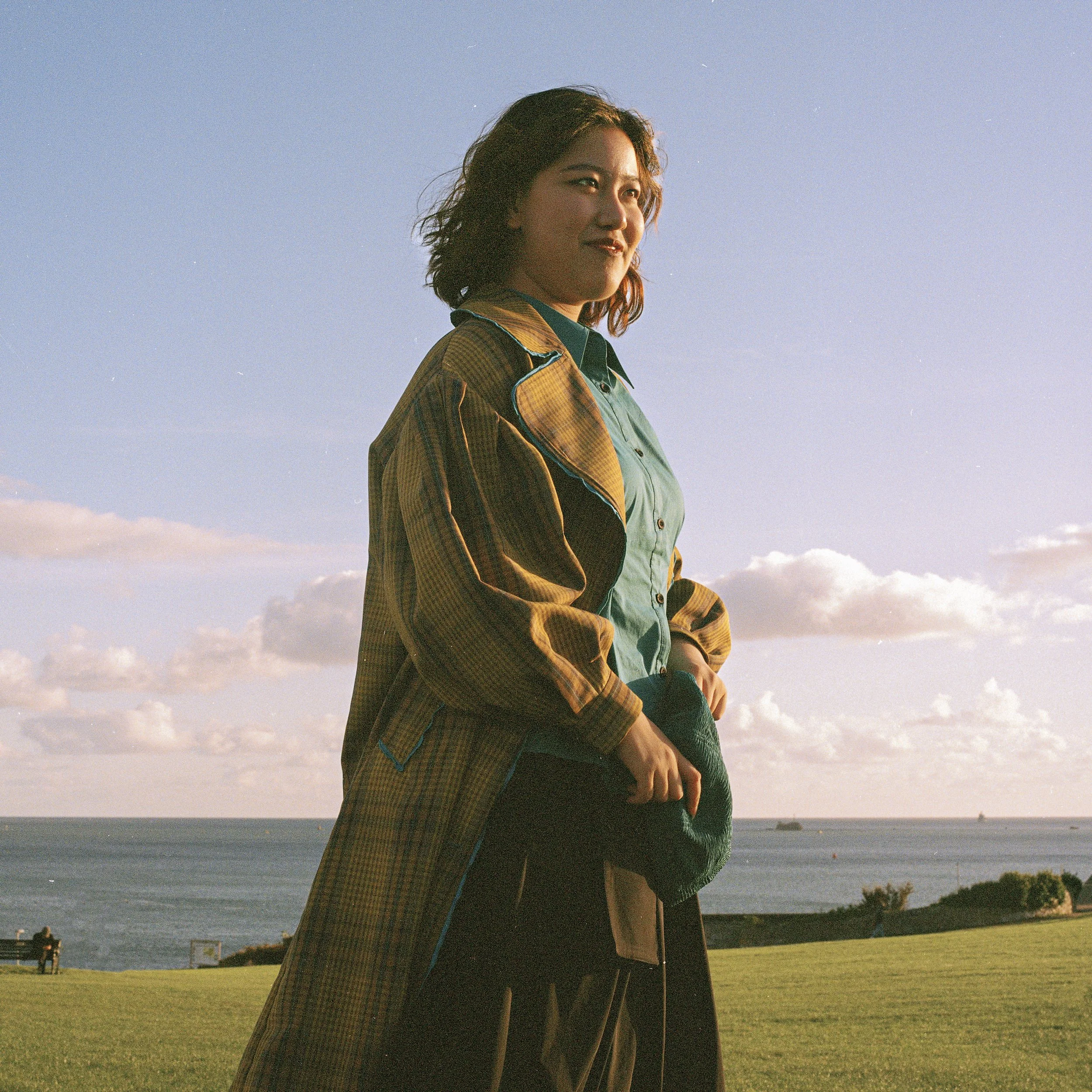 A woman standing outdoors on a grassy area near a body of water with trees in the background, during late afternoon or early evening, with a clear sky and some clouds.