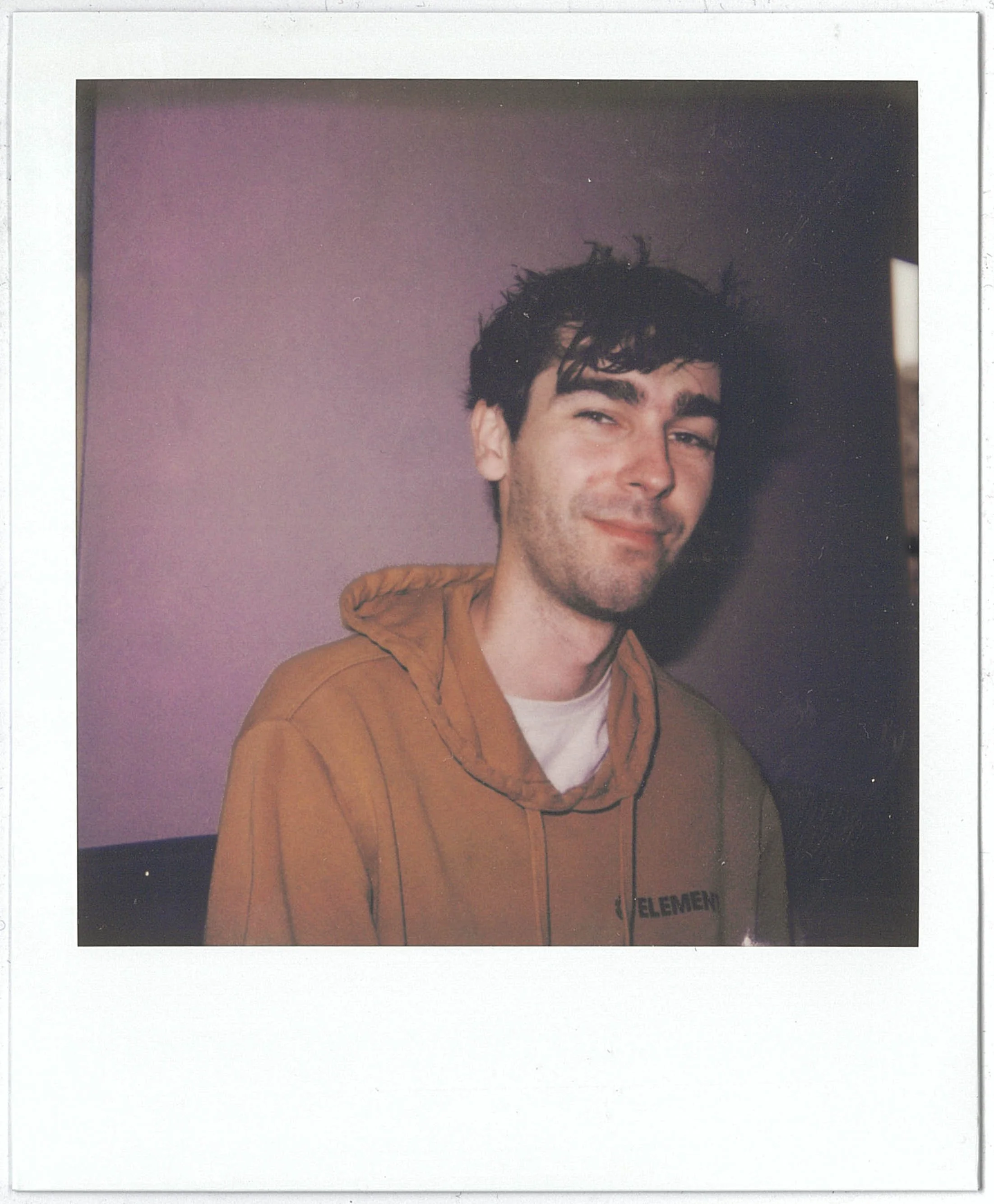 A Polaroid of a young man with dark, messy hair and a light beard, smiling softly, wearing a brown hoodie, standing against a purple wall.