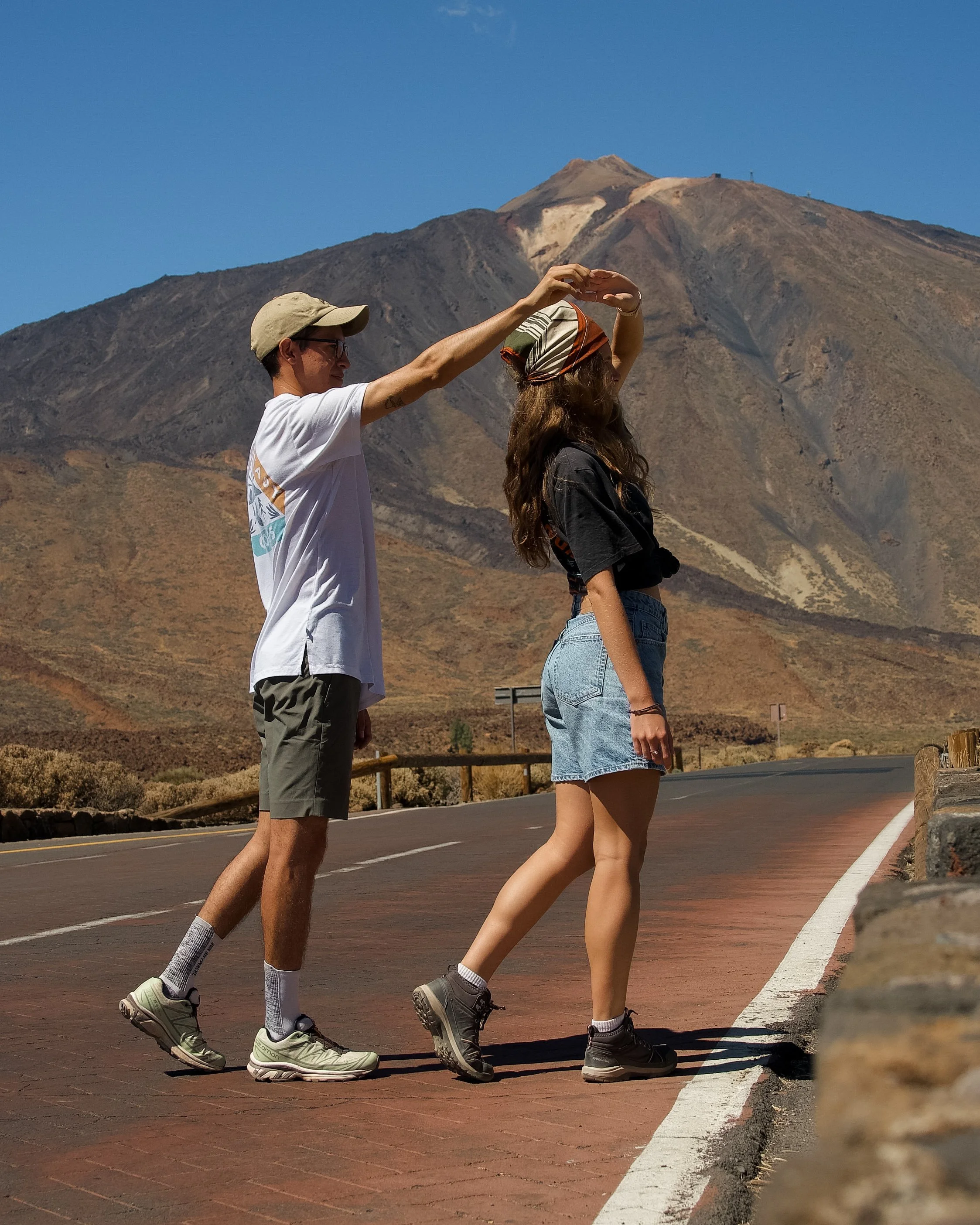 Dos personas caminando en la carretera con un volcán en el fondo, una de ellas ayudando a la otra a cruzar la línea blanca de la carretera.
