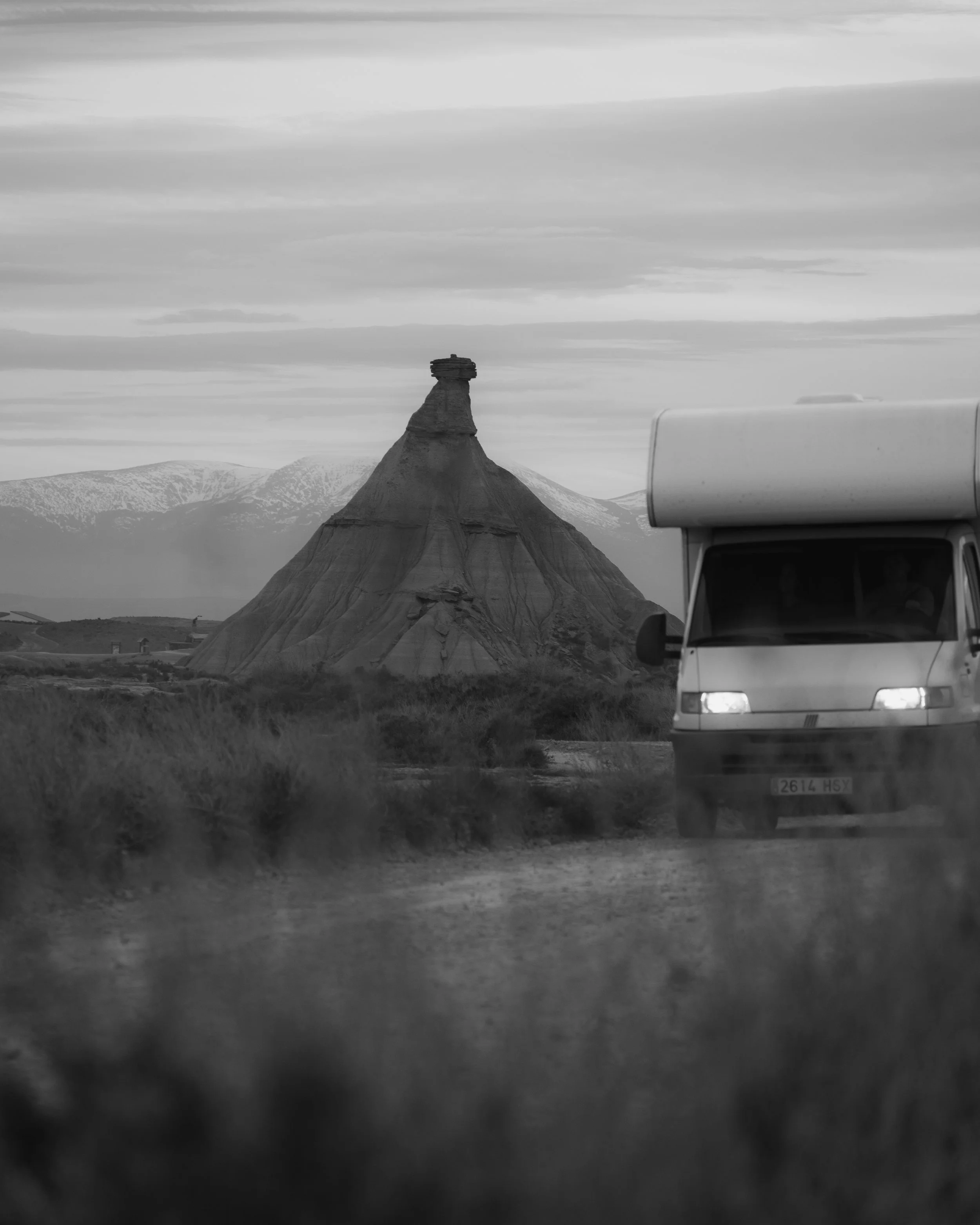 Vista en blanco y negro de un paisaje con una formación rocosa distintiva en el centro y un vehículo en primer plano a la derecha.