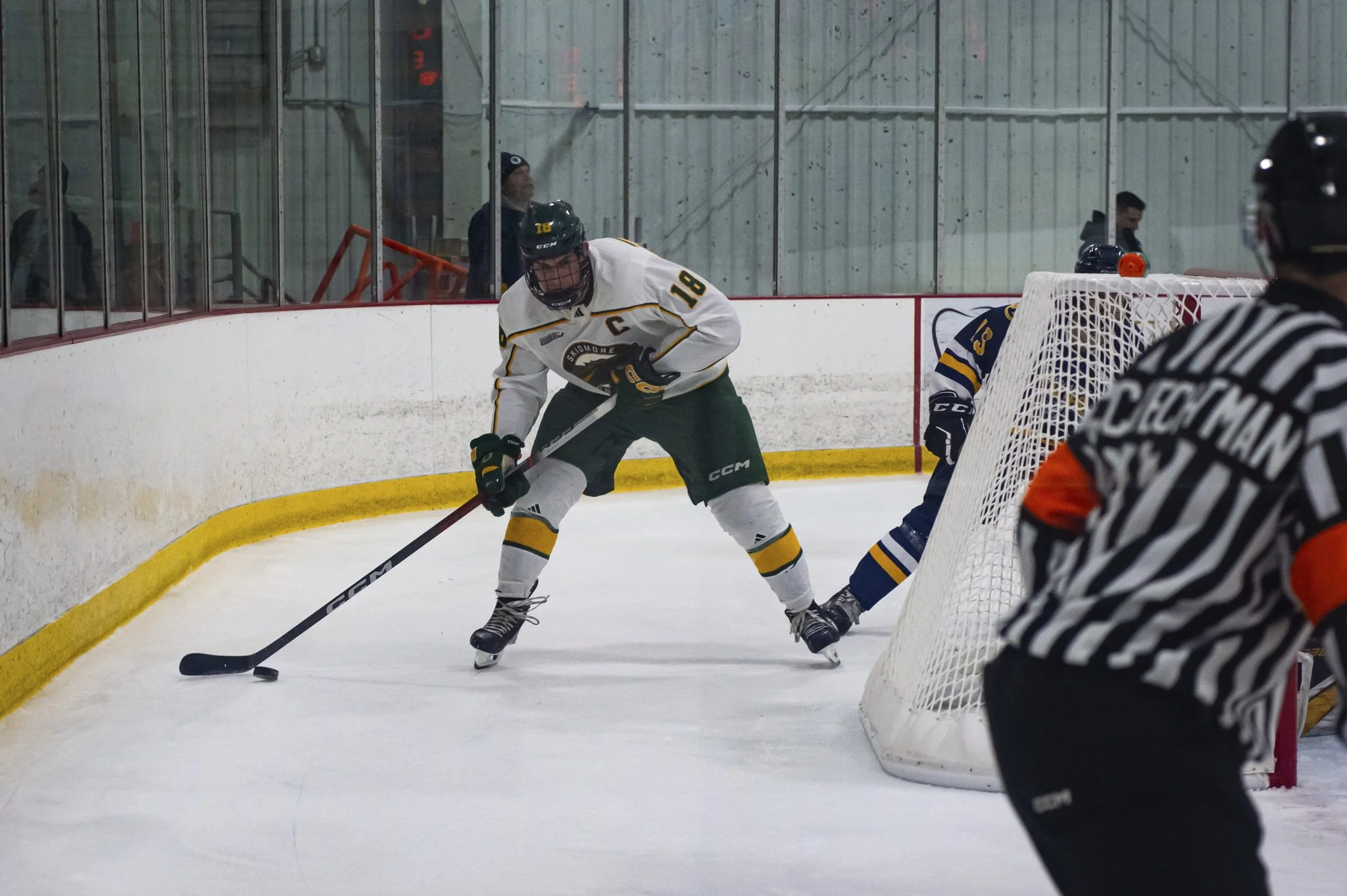 An ice hockey game with players in action near the goal, one player in a white and green uniform with the number 18 trying to control the puck, a player in a dark uniform with the number 16 standing behind, and an official in a black and white striped uniform nearby.
