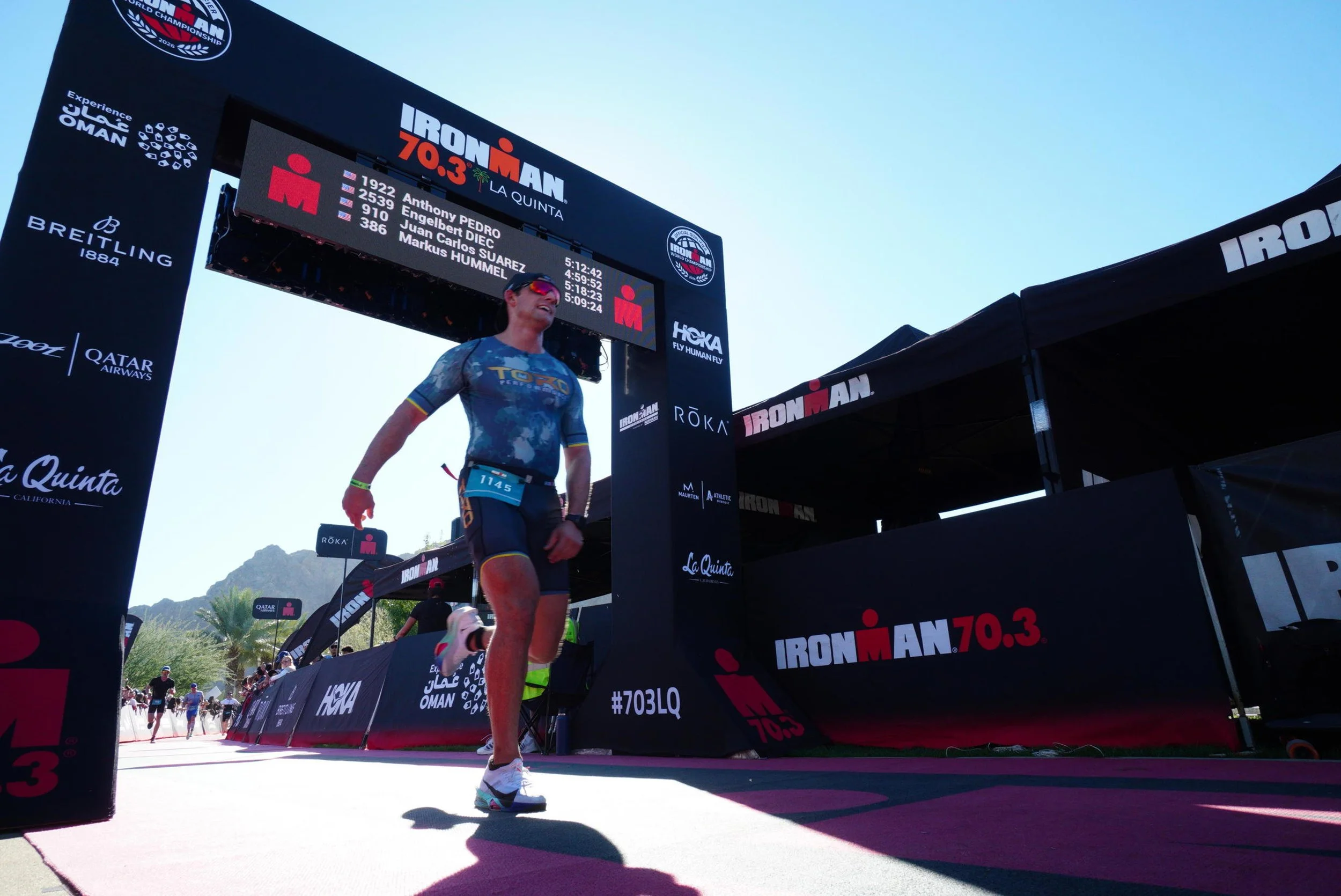 A male athlete crossing the finish line at the Ironman 70.3 triathlon event in La Quinta, California. The athlete is wearing sunglasses, a blue athletic suit, and a race bib. The background shows a bright, sunny day, with a digital leaderboard displaying race information.