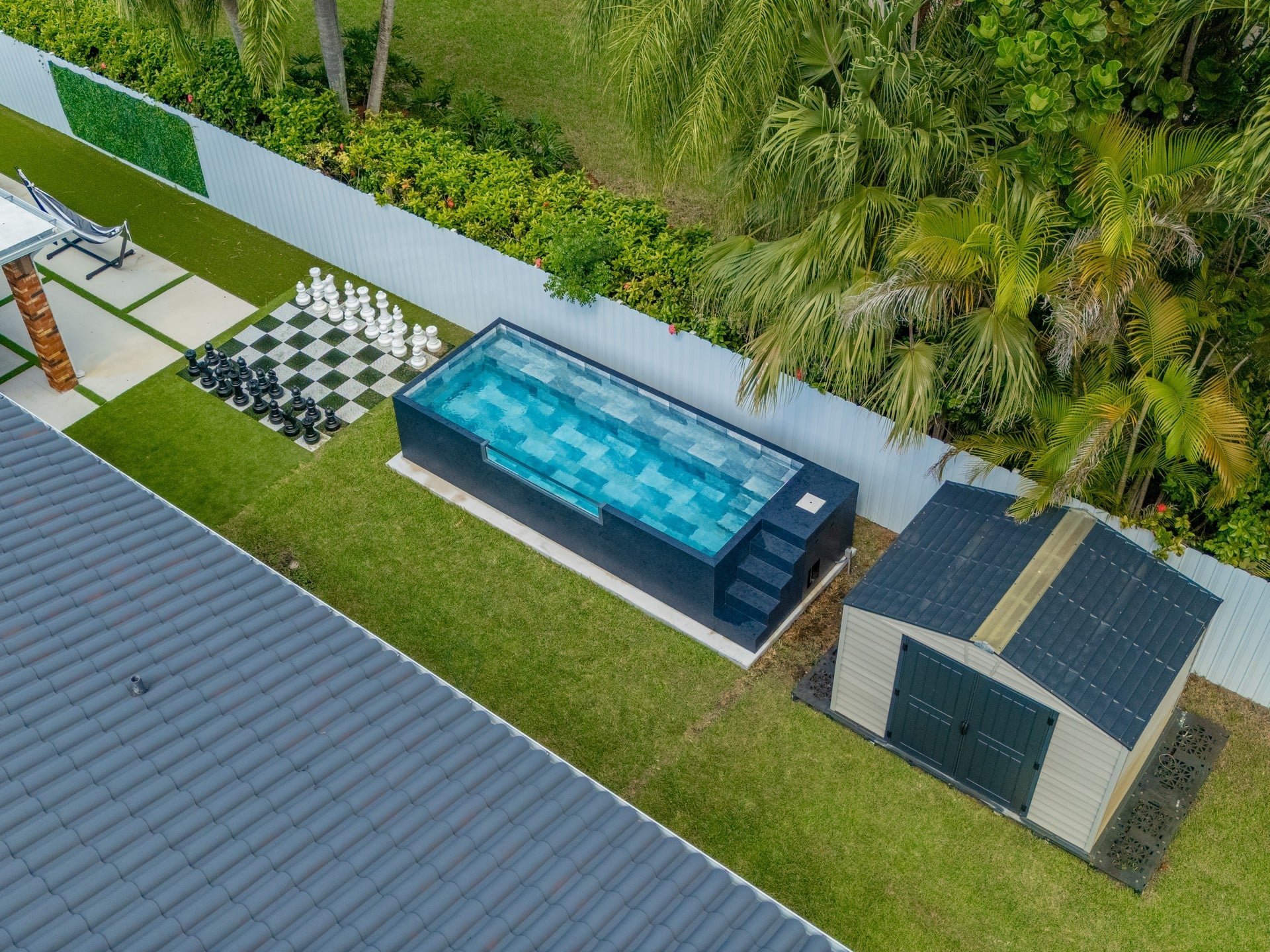 Aerial view of a courtyard with an above-ground pool, a giant chessboard, and a seating area with chairs, with green walls and lush vegetation in the background.