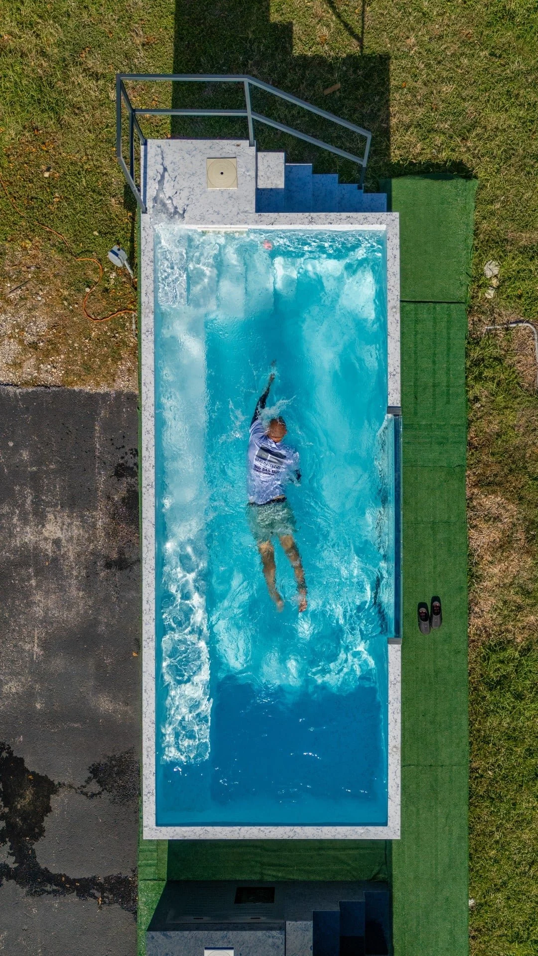 Aerial view of a rectangular above-ground pool with a person swimming in it, surrounded by grass and paved areas, with stairs at one end and a shadow cast over the top.