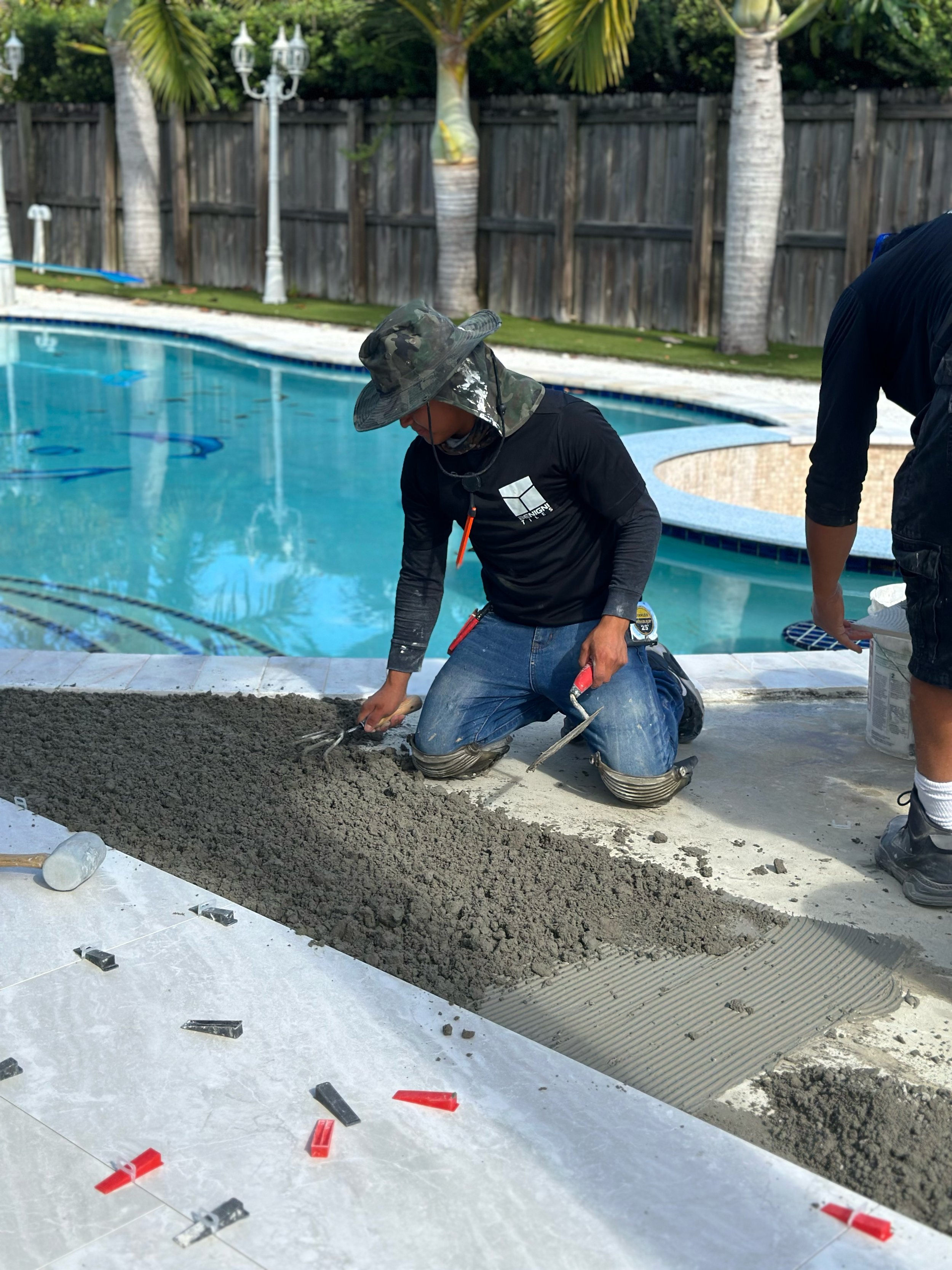 A worker laying tiles for a pool, with tools and construction materials, in a landscaped area with palm trees and near a wooden fence.