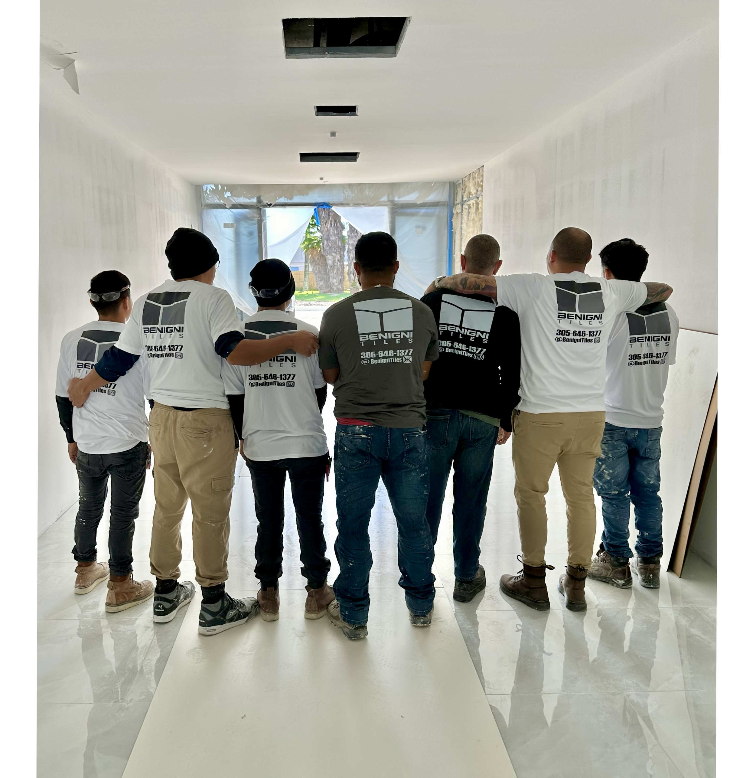A group of six people with their backs to the camera in a white hallway, some wearing white T-shirts with the Benigni Tiles logo and a contact number, at some kind of meeting or event