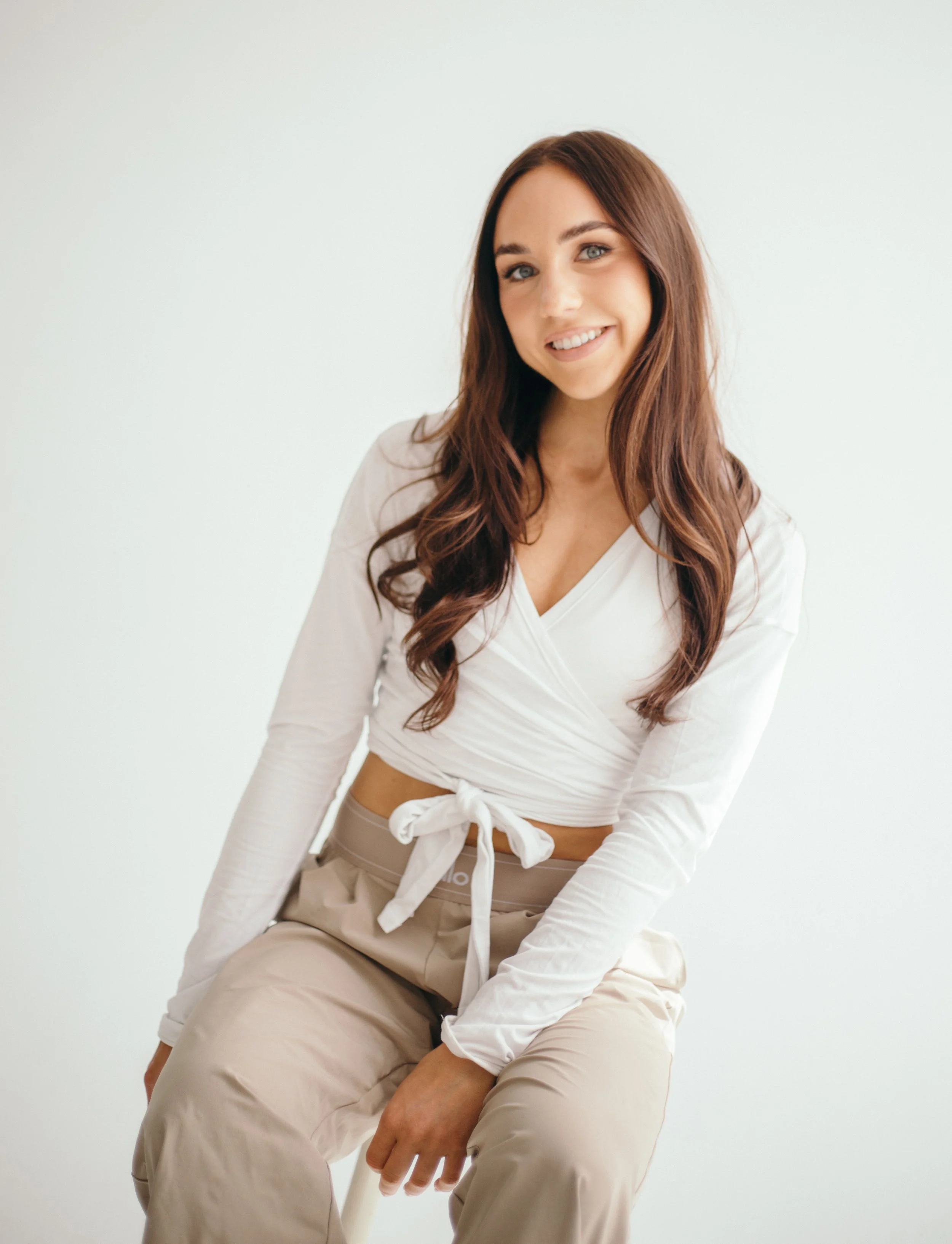 A young woman with long brown hair, wearing a white wrap top and beige pants, smiling softly and sitting against a plain white background.