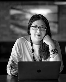 Woman with glasses sitting at a table with an open laptop, smiling and resting her chin on her hand in a modern indoor setting.
