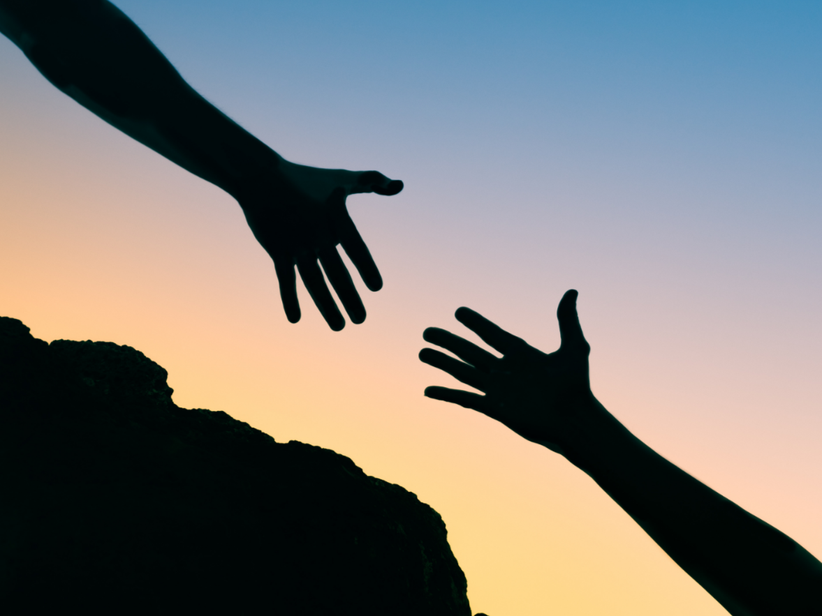 Two silhouetted hands reaching toward each other against a sunset sky, one hand on a rock or cliff edge.