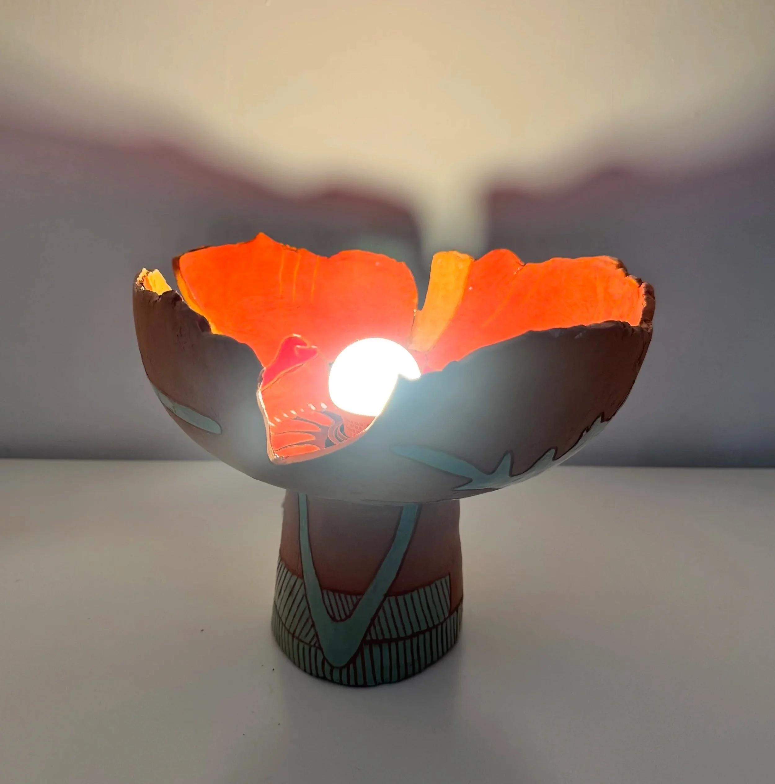 A decorative poppy inspired lamp with a colorful, abstract pattern and a glowing light bulb inside, placed on a white surface against a plain background.