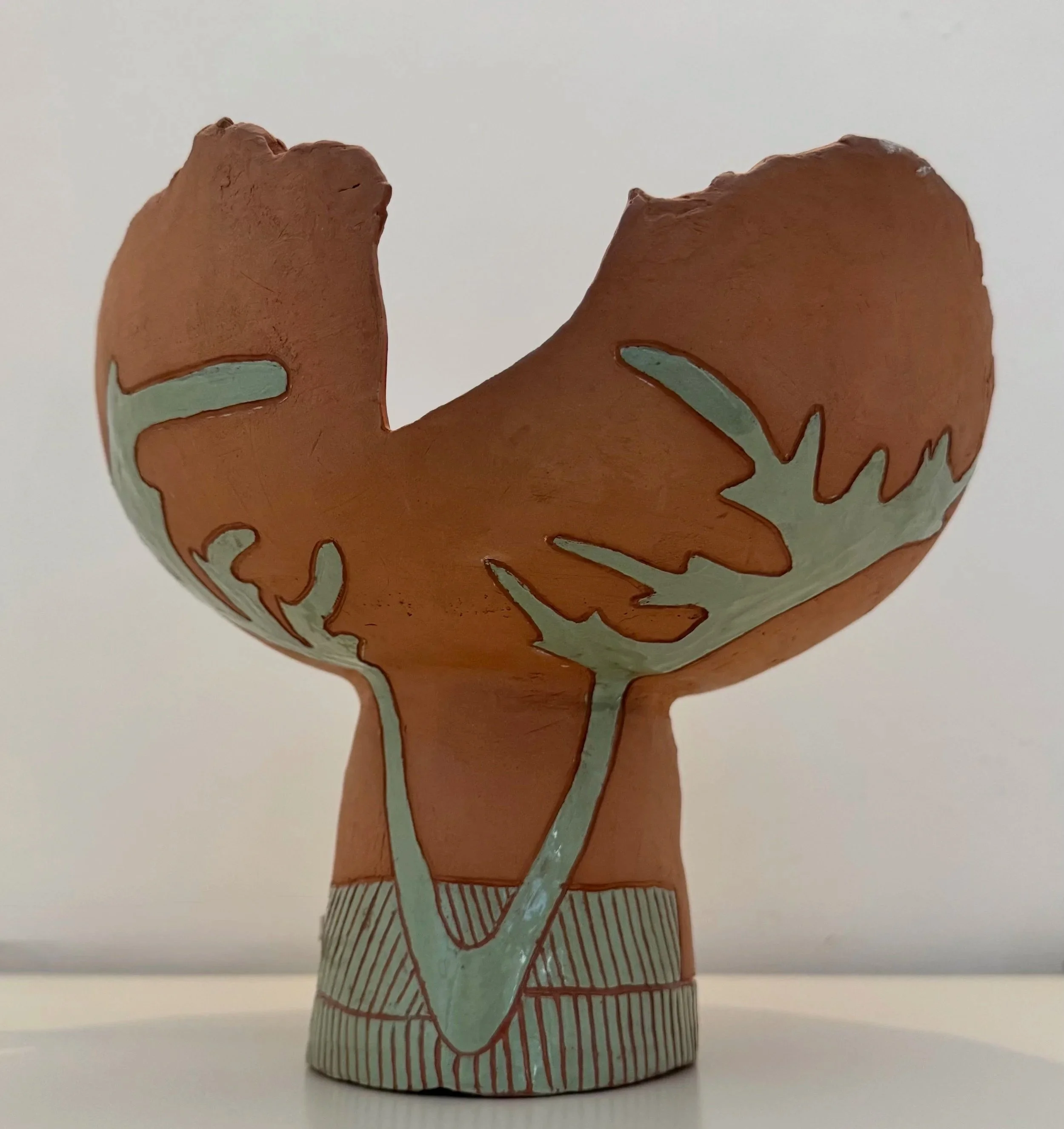 A ceramic sculpture of a bear's head and upper body, painted in earthy brown and green colors, with decorative line patterns, set against a plain white background.